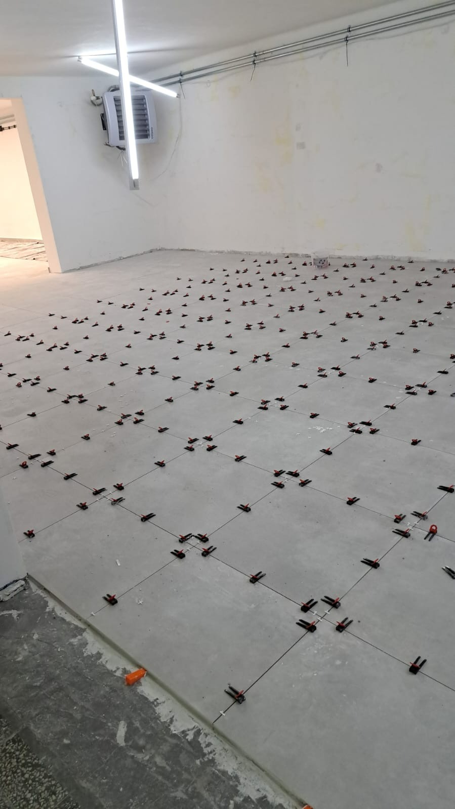 Układanie płytek podłogowych w dużym pomieszczeniu, widoczne krzyżyki dystansowe i system poziomowania płytek, surowy stan wykończenia ścian i podłogi.