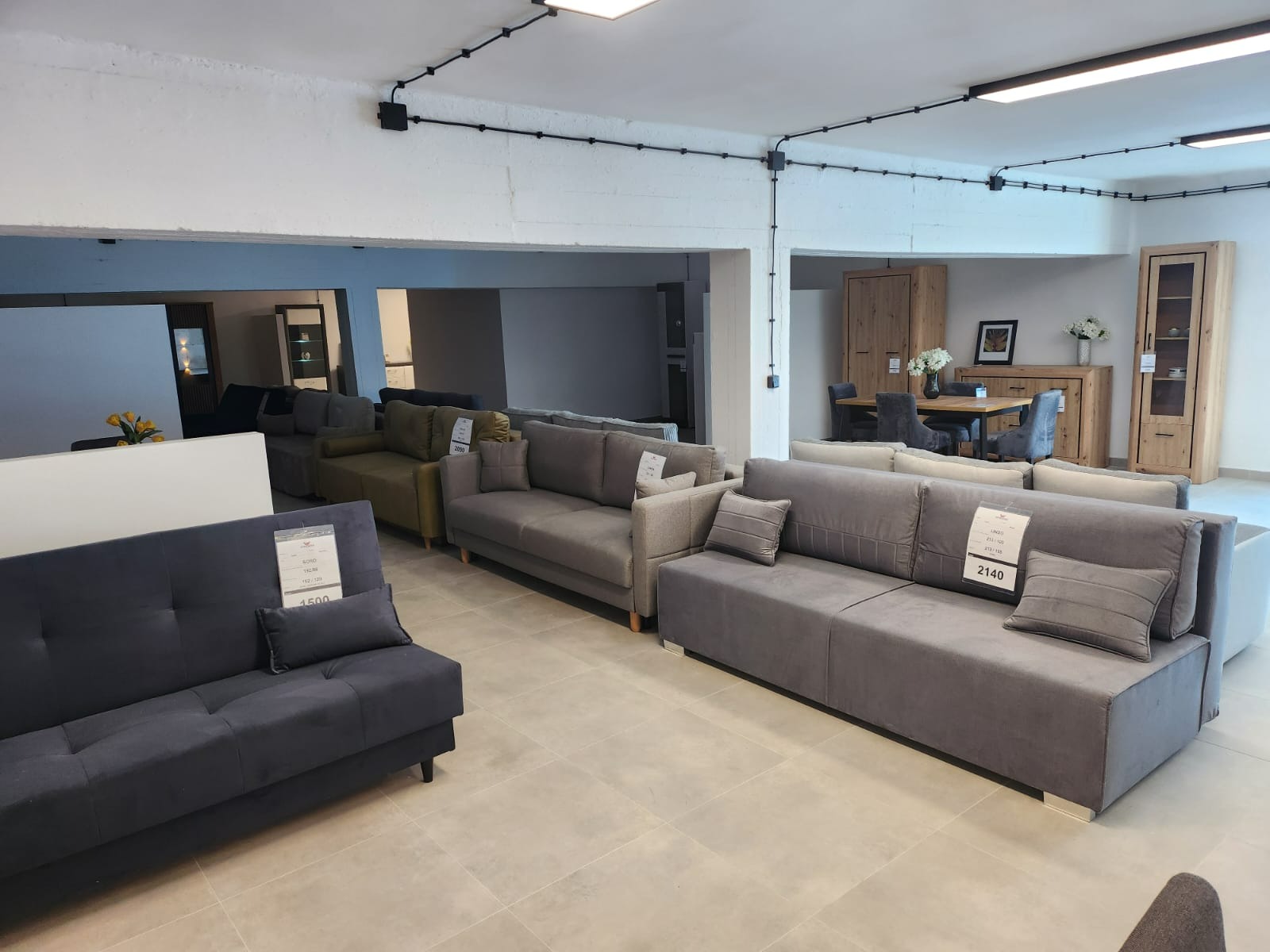 Ekspozycja w salonie meblowym: Rzędy nowoczesnych sof w odcieniach szarości i oliwki, z poduszkami i cenami, na jasnej podłodze z płytek, w tle aranżacja jadalni z drewnianymi meblami.