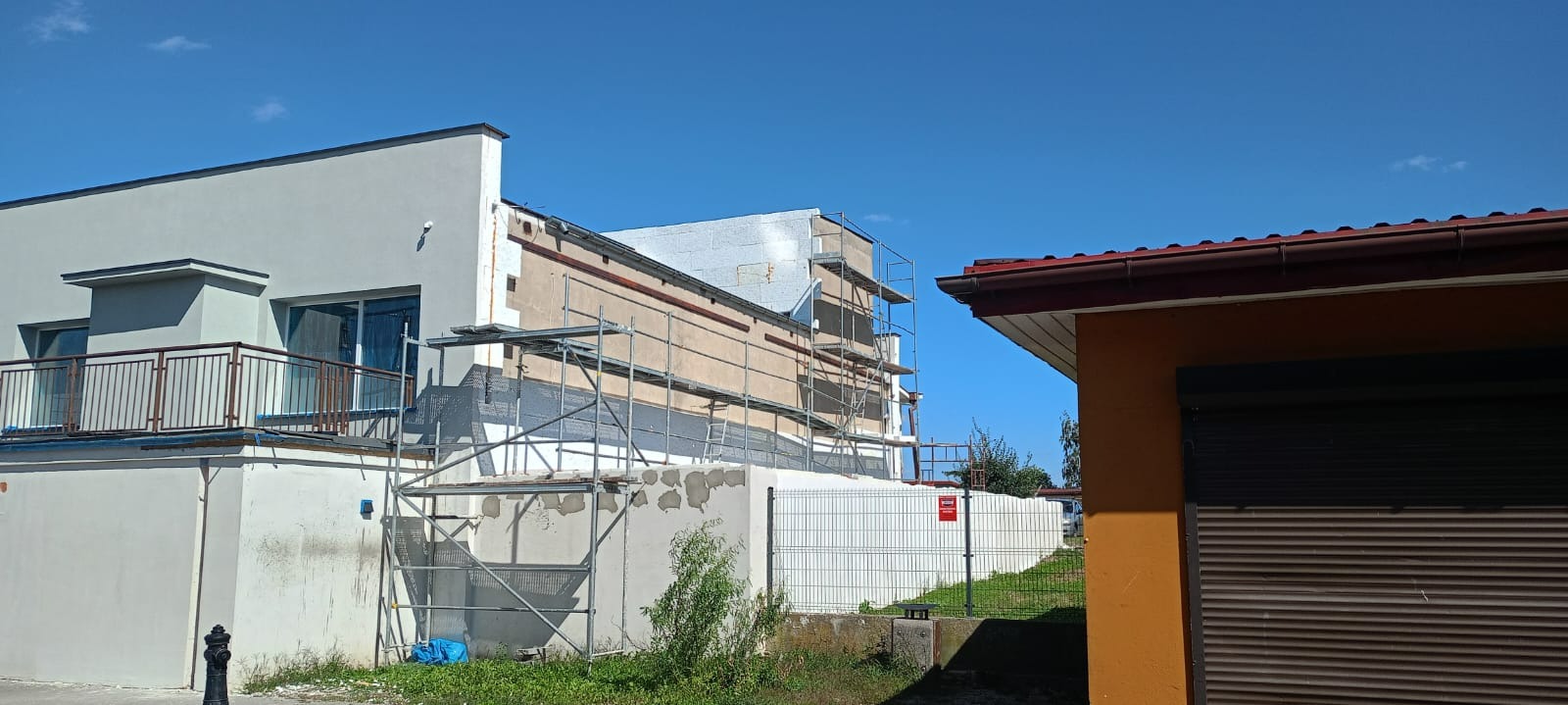 Budynek w trakcie prac elewacyjnych: fragment ocieplonej ściany, rusztowania, oraz fragment budynku po tynkowaniu i malowaniu na szaro, błękitne niebo w tle.