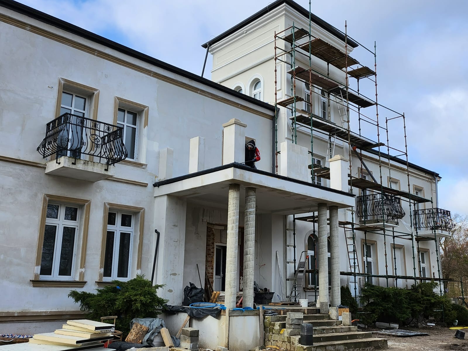 Prace wykończeniowe elewacji budynku z balkonami i kolumnami, widoczne rusztowania i pracownik w trakcie wykonywania poprawek.