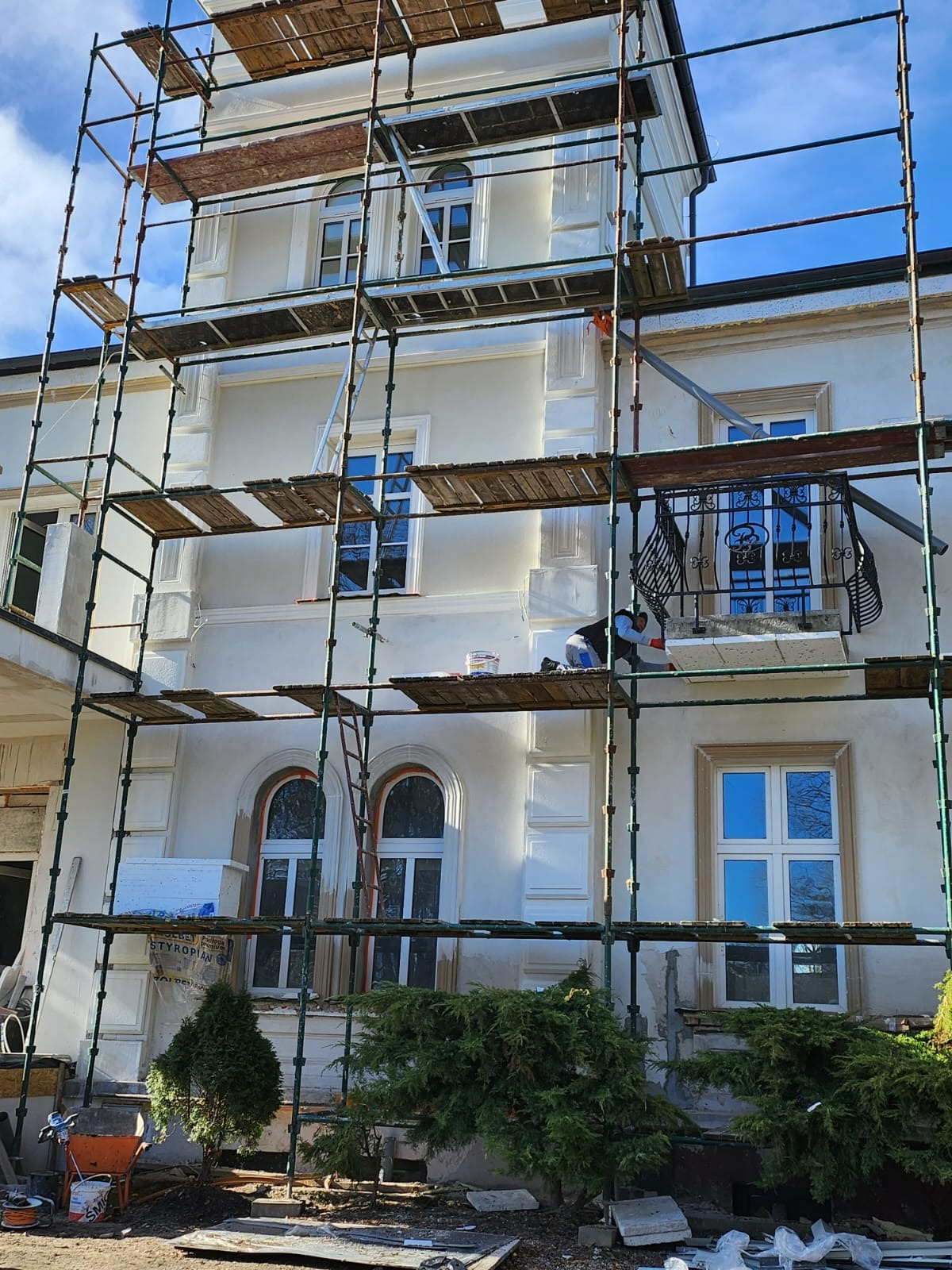Elewacja budynku w trakcie prac wykończeniowych, widoczne rusztowania, pracownik na rusztowaniu przy balkonie, styropianowe płyty i narzędzia na ziemi.