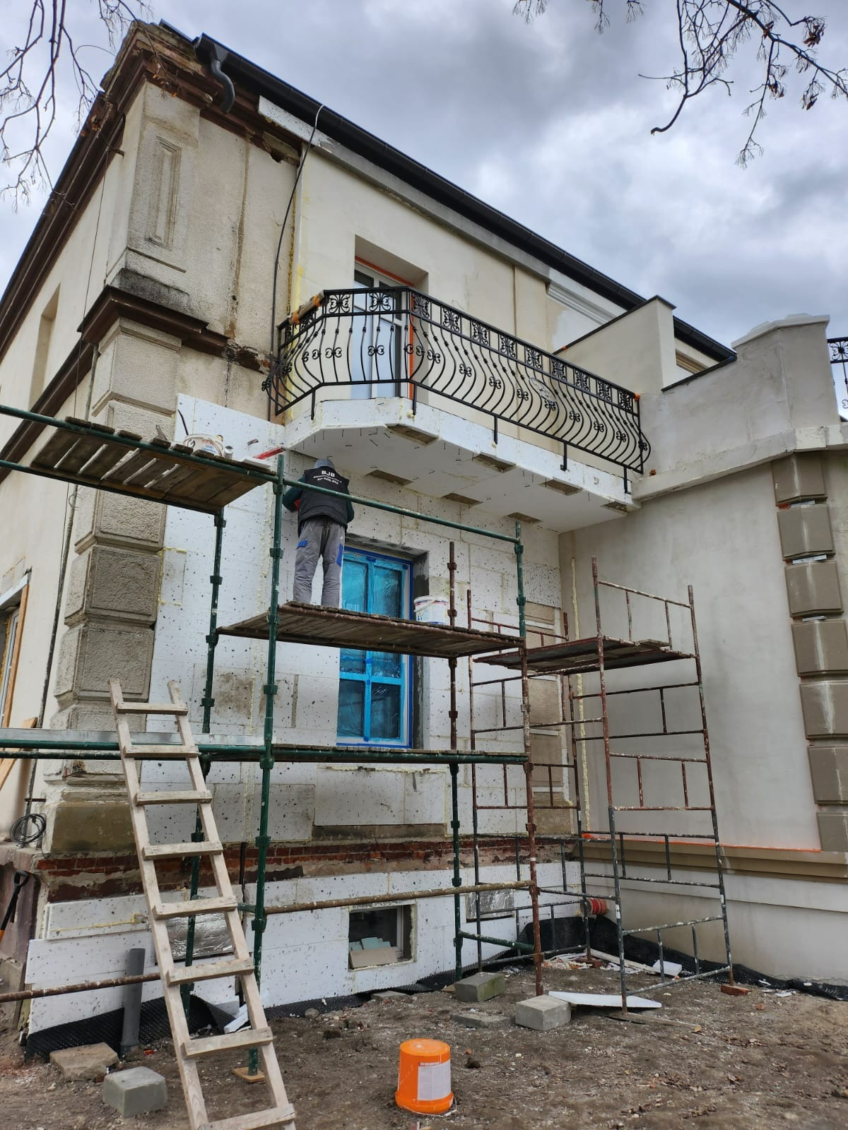 Pracownik na rusztowaniu nakłada warstwę izolacyjną na elewację budynku z balkonem, okno zabezpieczone folią, widoczne fragmenty starej i nowej elewacji.