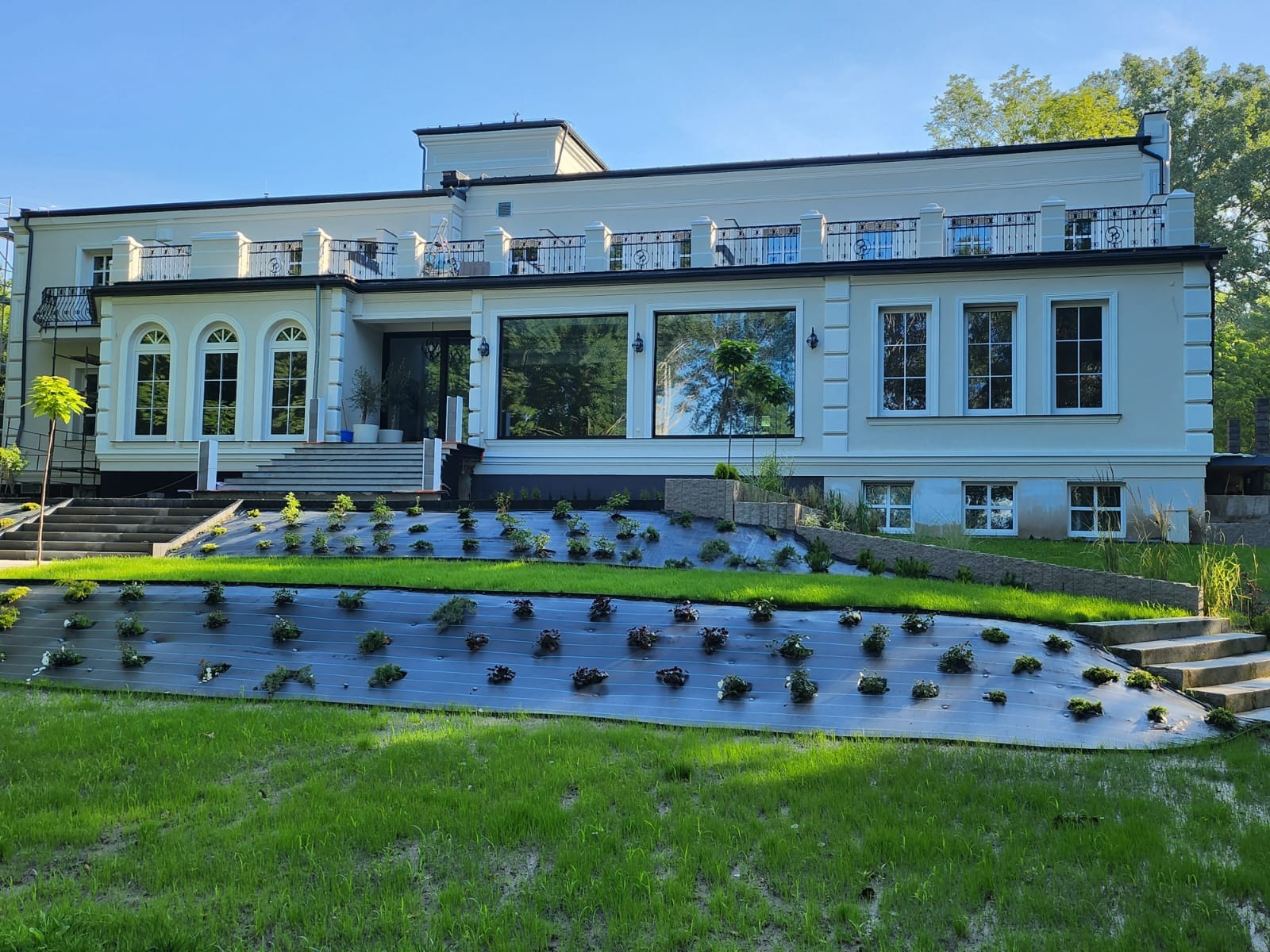 Jasny, dwukondygnacyjny dom z balkonami i dużymi oknami, z zagospodarowanym terenem przed budynkiem, gdzie widoczna jest agrowłóknina z nasadzeniami.