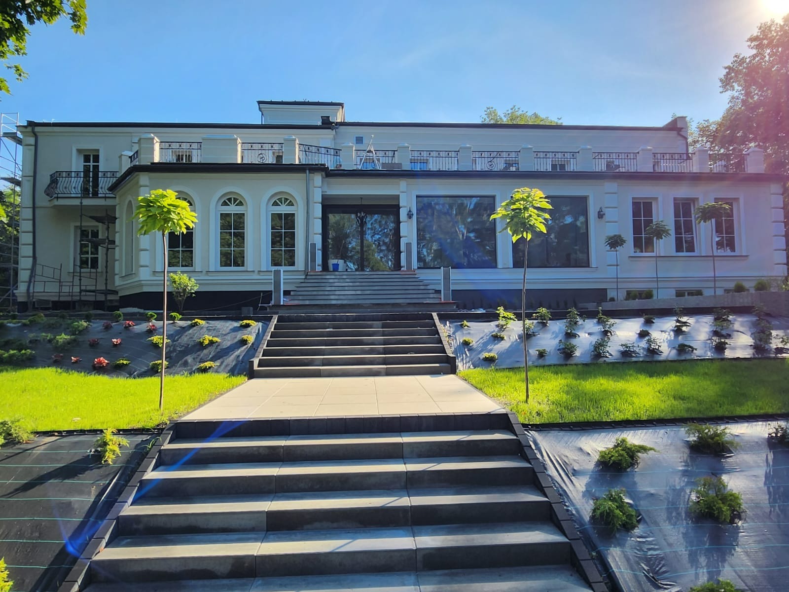 Jasny, dwupiętrowy budynek z balkonami i dużymi oknami, widoczny od frontu z perspektywy schodów prowadzących do wejścia, otoczony świeżo założonym trawnikiem i młodymi drzewkami.