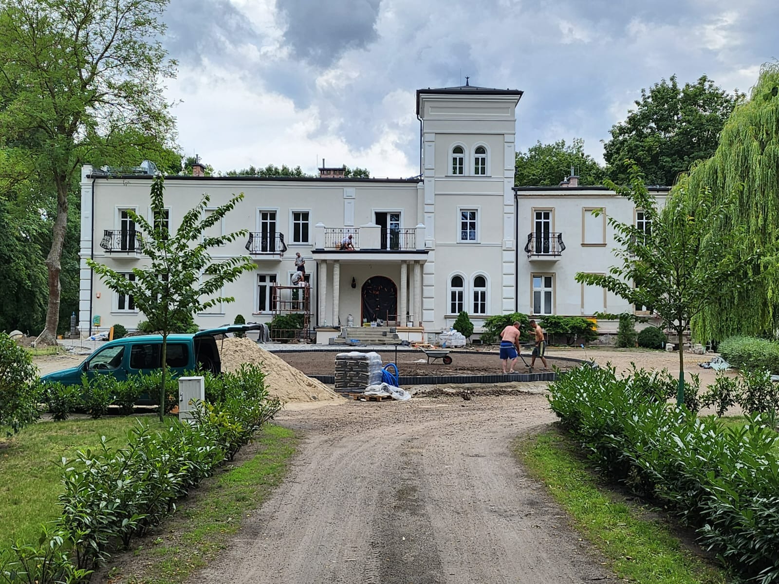 Dwupiętrowy budynek z wieżą w trakcie prac elewacyjnych, widoczne rusztowania i pracownicy. Na pierwszym planie przygotowanie terenu pod kostkę brukową, sterta piasku i otwarty samochód dostawczy.
