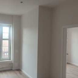 REMOLUKI ŁUKASZ LITWIŃSKI - Jasne wnętrze po odświeżeniu: pomalowane ściany w odcieniach bieli, drewnopodobna podłoga, białe okno z widokiem na budynki, minimalistyczna lampa ścienna i widoczne ślady po starym grzejniku...