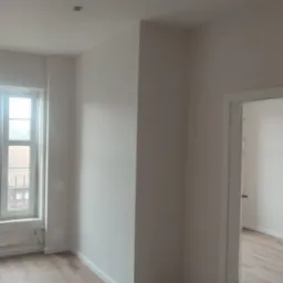 Jasne wnętrze po odświeżeniu: pomalowane ściany w odcieniach bieli, drewnopodobna podłoga, białe okno z widokiem na budynki, minimalistyczna lampa ścienna i widoczne ślady po starym grzejniku...