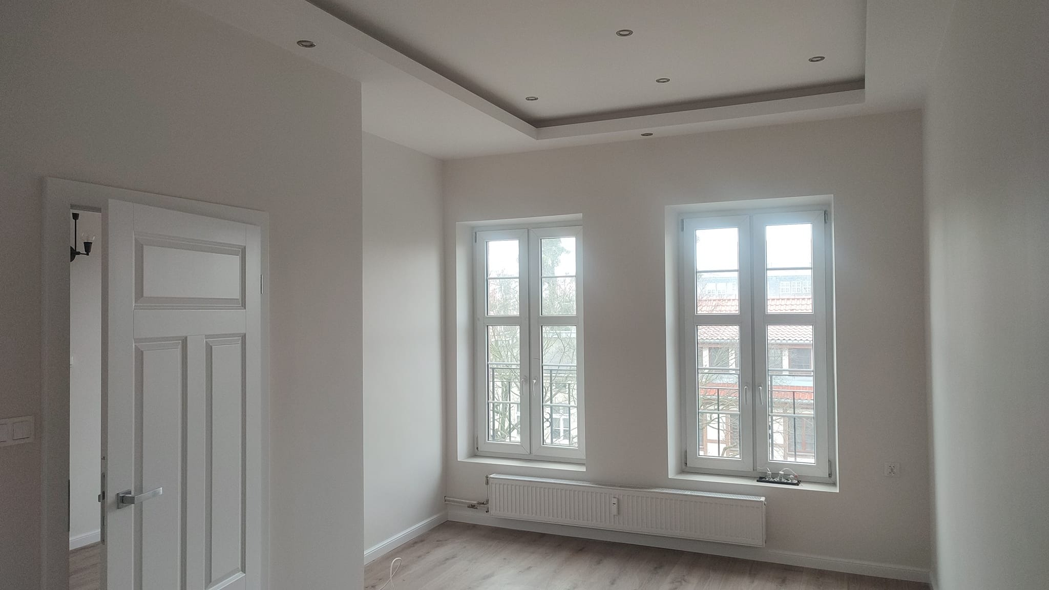 Pomalowane na biało wnętrze z dwoma oknami, grzejnikiem i drzwiami z panelami, jasna podłoga, sufit z podwieszaną częścią i oświetleniem punktowym.