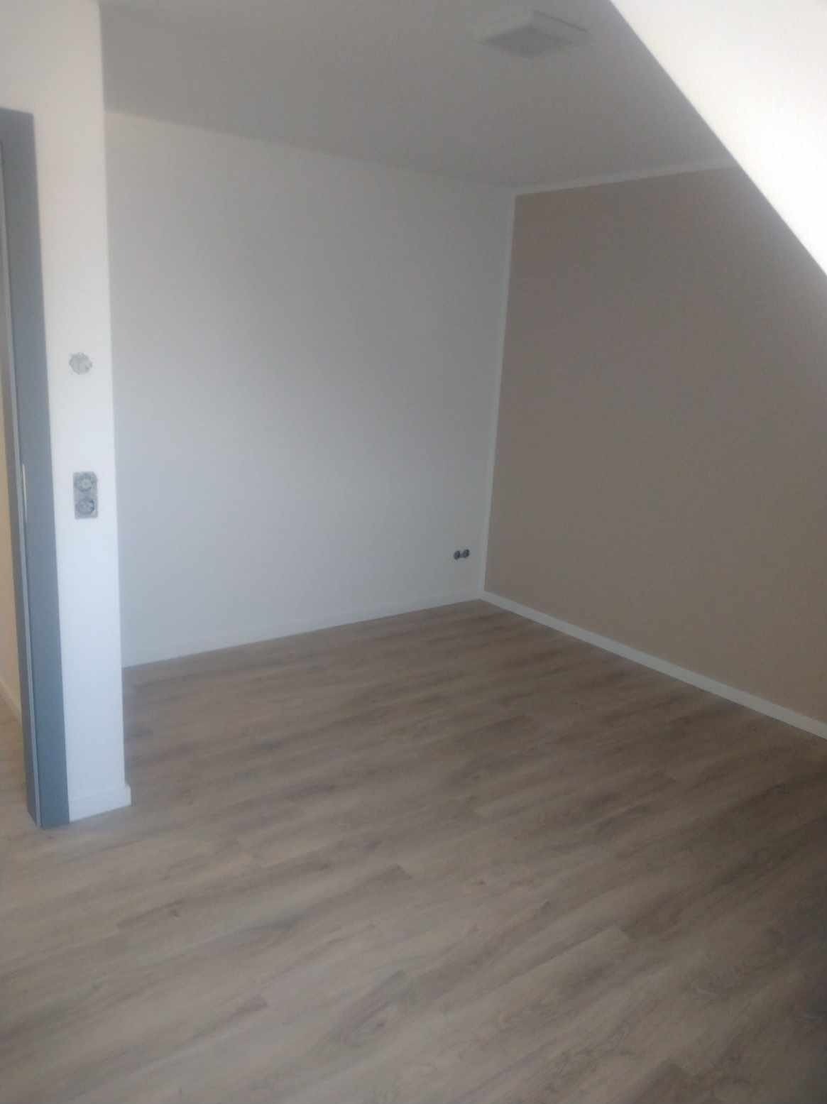 Pomalowane wnętrze poddasza z nową podłogą, ściany w odcieniach bieli i beżu, widoczne gniazdka elektryczne i listwa przypodłogowa.