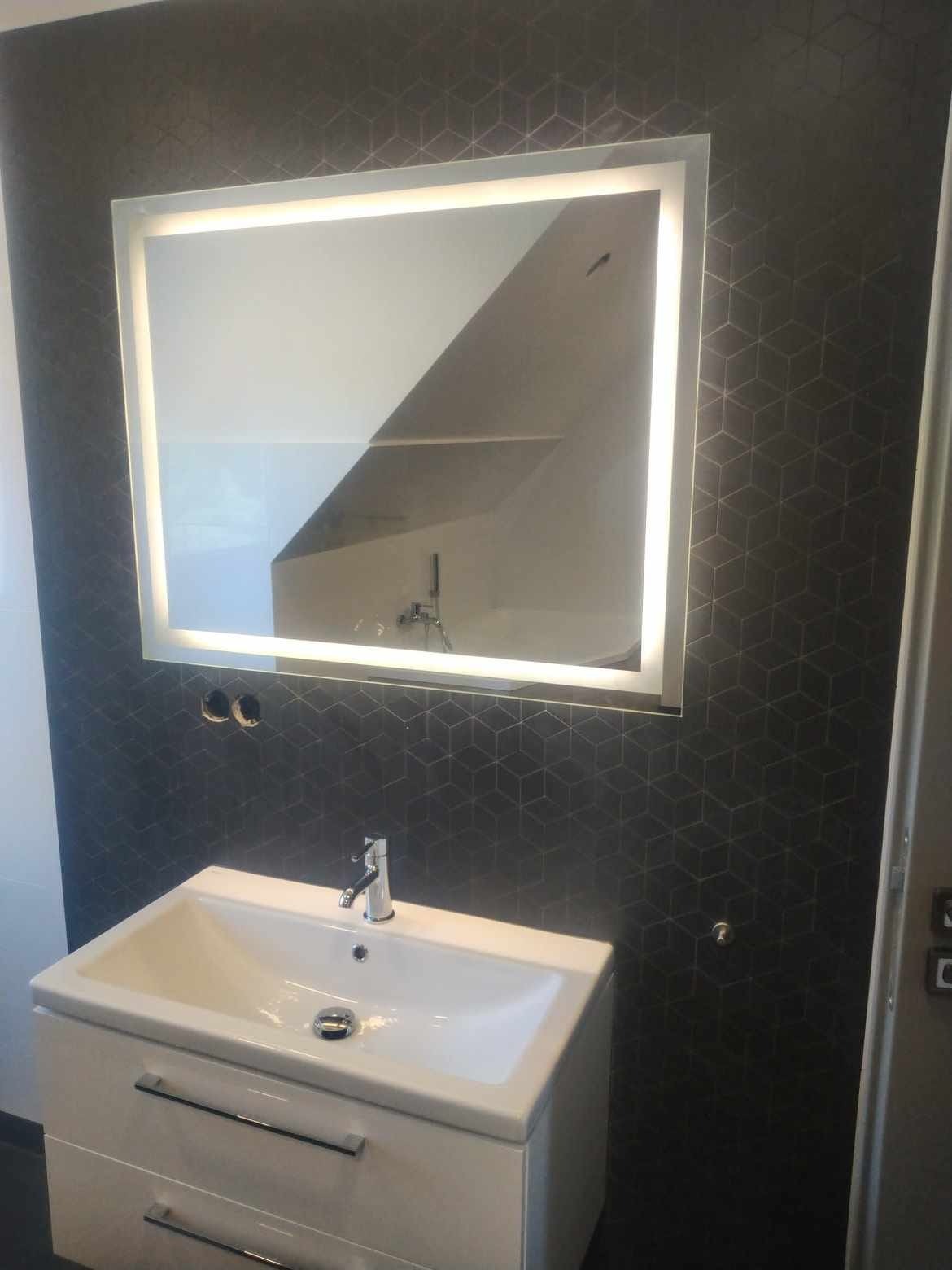Nowoczesna łazienka z umywalką na szafce, podświetlanym lustrem i ciemną ścianą w geometryczny wzór; widoczne gniazdka elektryczne i chromowana bateria.