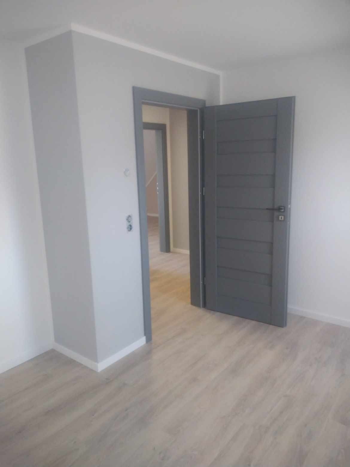 Szare, panelowe drzwi wewnętrzne z ościeżnicą w odcieniu szarości, uchylone w nowo wyremontowanym pokoju z jasnymi ścianami i panelami podłogowymi imitującymi drewno, widok na kolejne pomieszczenie...