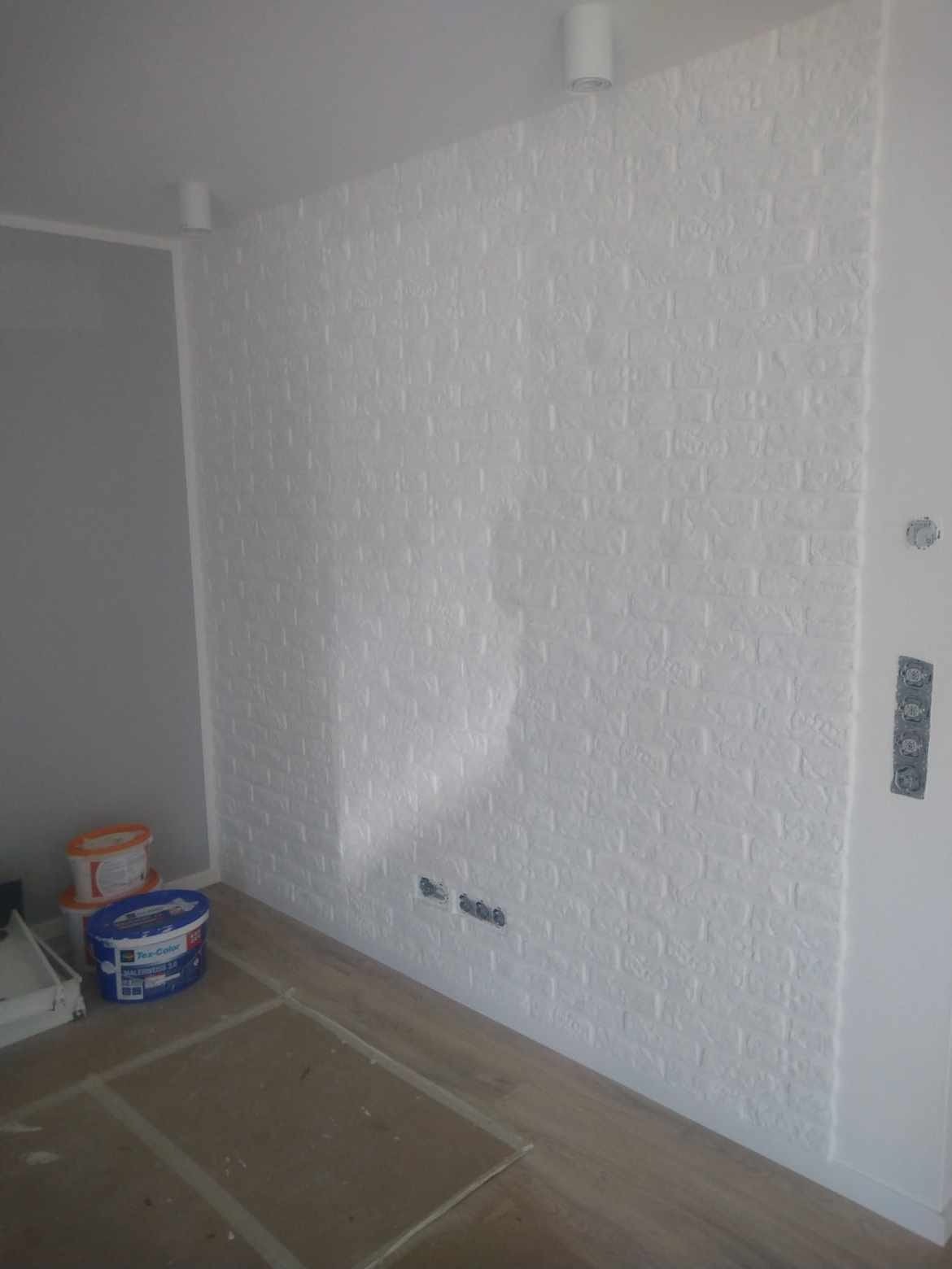 Ściana w pokoju pokryta białą farbą imitującą cegłę, widoczne puszki z farbą i zaklejona podłoga.