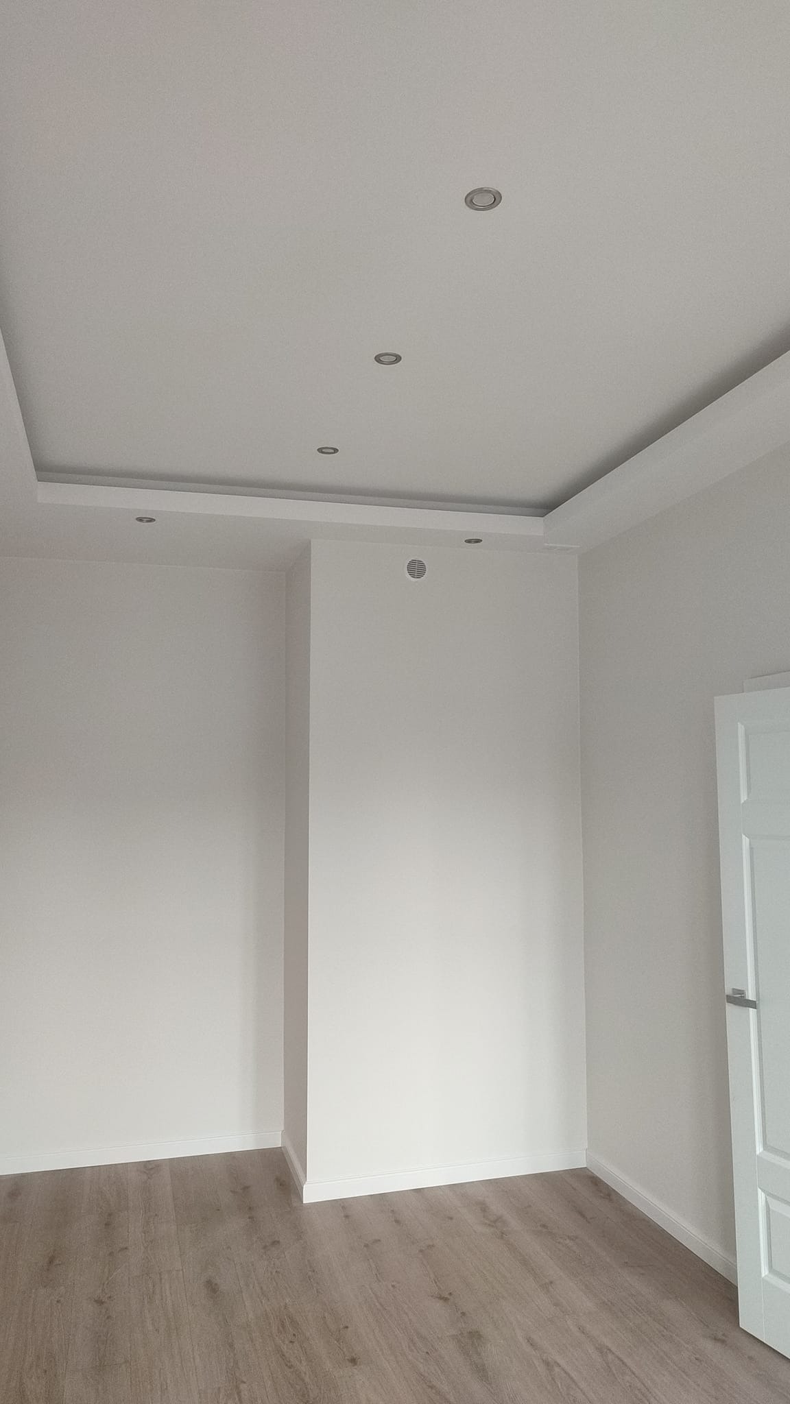 Jasne, nowocześnie wykończone wnętrze z drewnianą podłogą, białymi ścianami, ozdobnym sufitem podwieszanym z oświetleniem punktowym i fragmentem białych drzwi po prawej stronie.