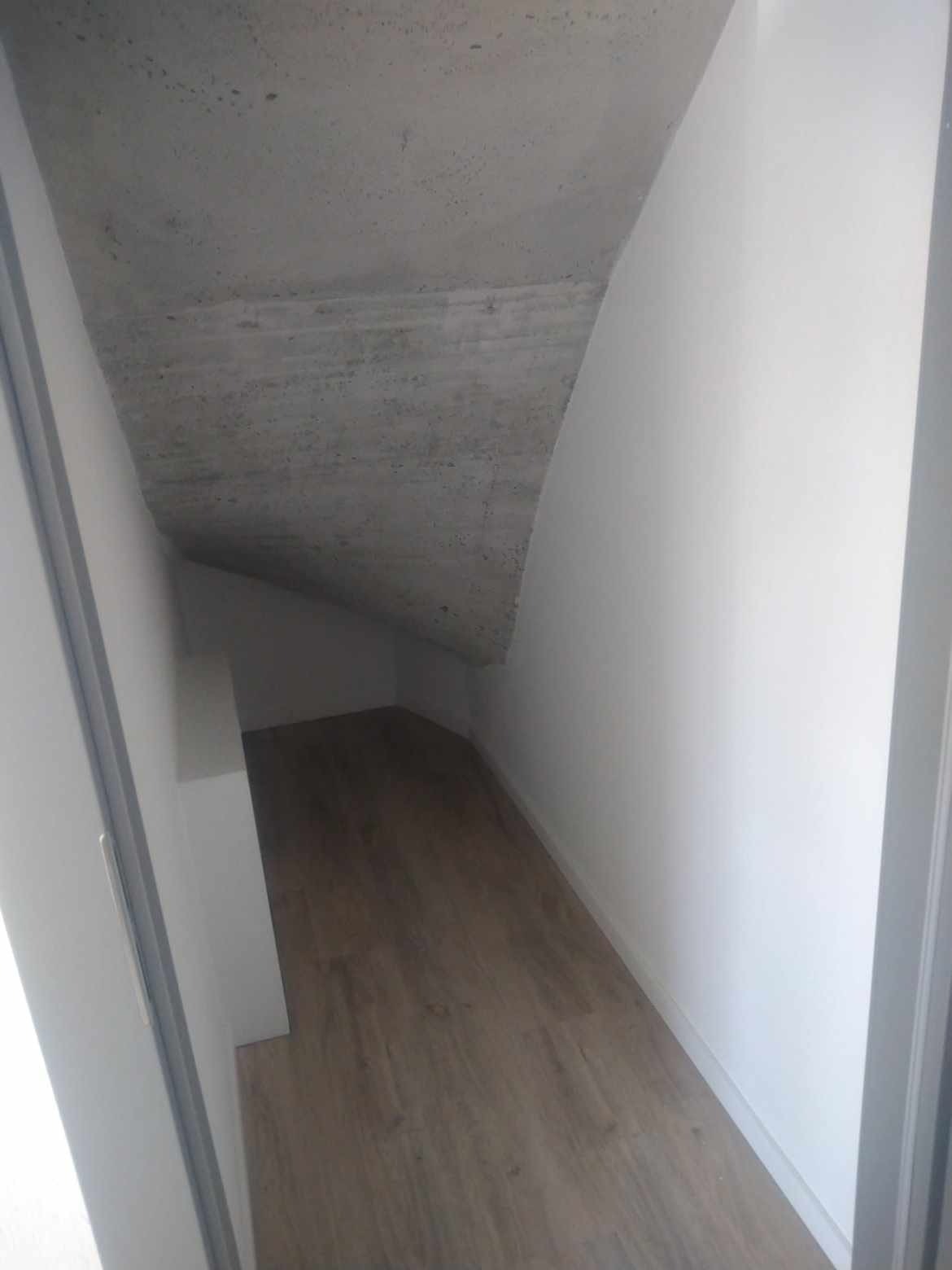 Wykończone wnętrze: skośny strych z podłogą z paneli i białą ścianą, widoczny surowy betonowy strop.