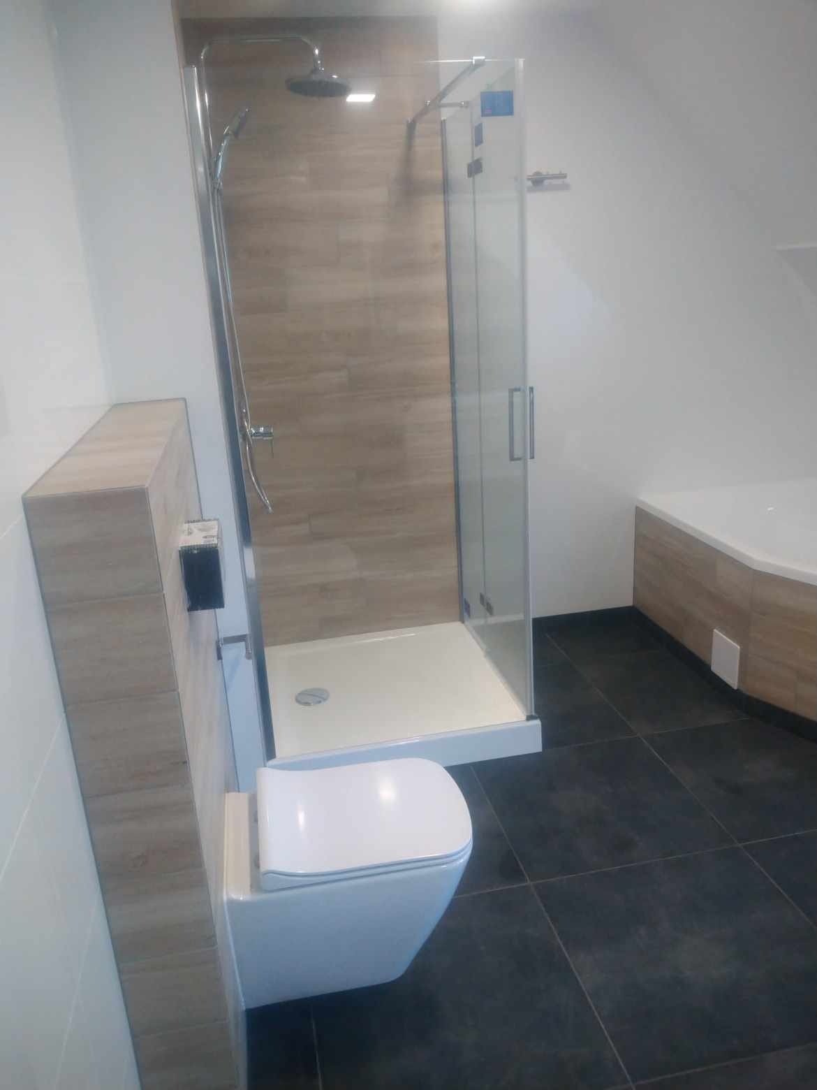 Nowoczesna łazienka z kabiną prysznicową typu walk-in z drewnopodobnymi płytkami na ścianie, podwieszaną toaletą i ciemną podłogą z płytek. Widoczny fragment wanny z obudową w tym samym stylu co...