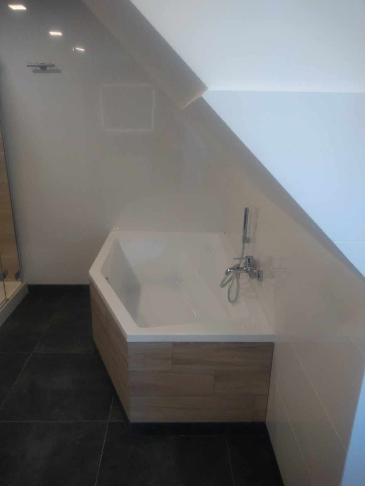 Wykończona łazienka na poddaszu ze skośnym sufitem, prezentująca białą, sześciokątną wannę obudowaną panelami imitującymi drewno, czarne płytki podłogowe i chromowaną armaturę.