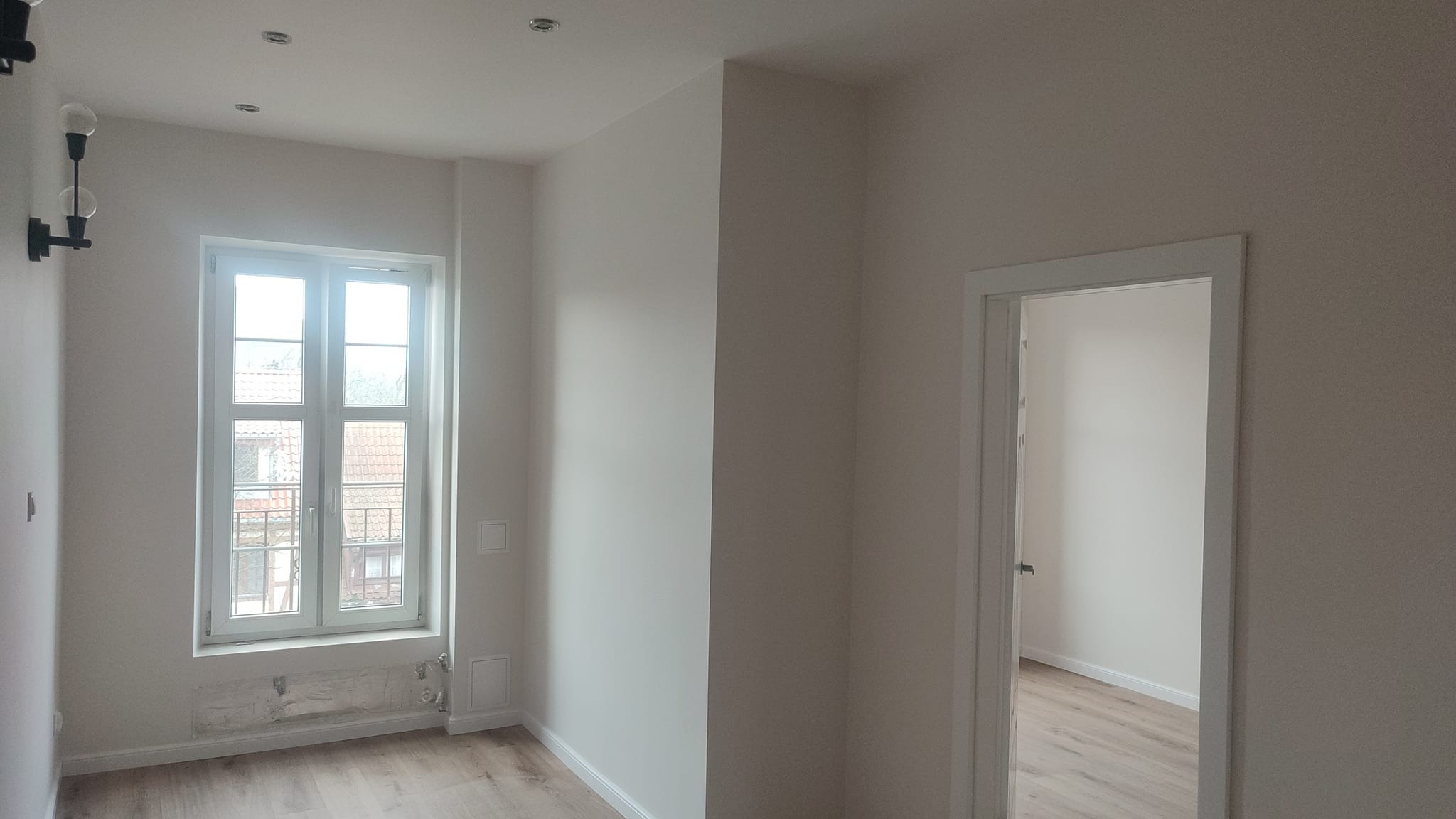 Jasne wnętrze po odświeżeniu: pomalowane ściany w odcieniach bieli, drewnopodobna podłoga, białe okno z widokiem na budynki, minimalistyczna lampa ścienna i widoczne ślady po starym grzejniku...