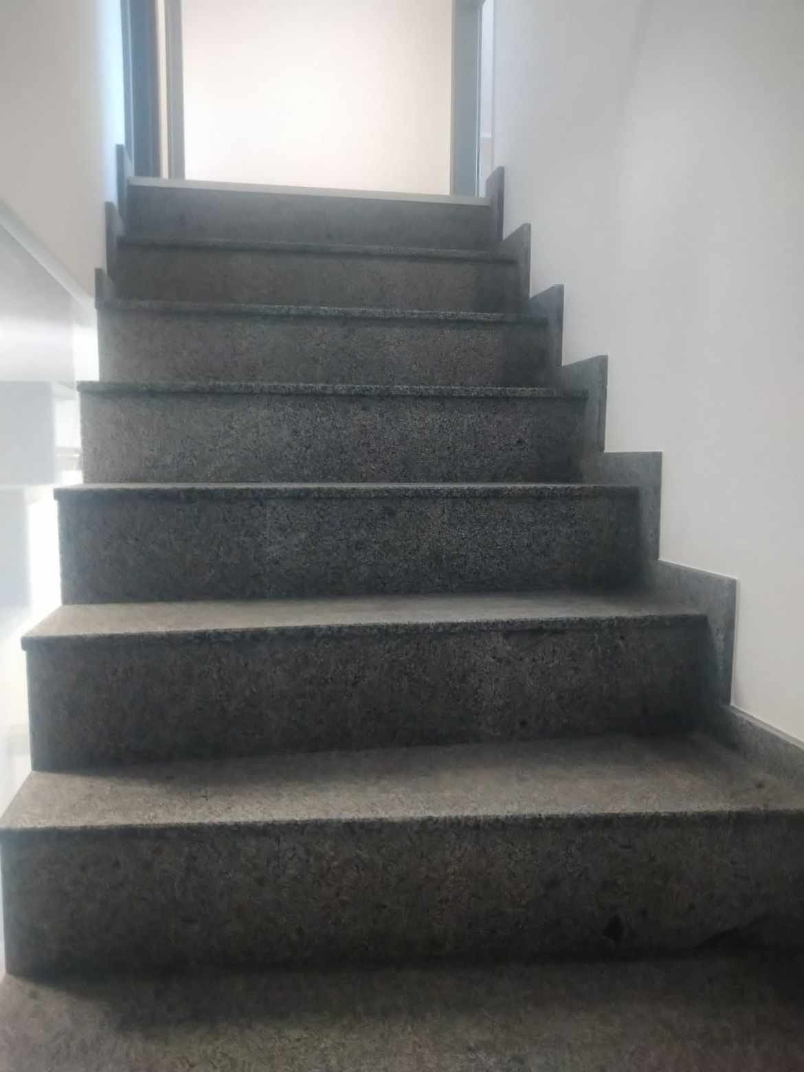 Granitowe schody wewnętrzne o szorstkiej fakturze, widok od dołu, z białymi ścianami w tle i jasnym przejściem na górze.