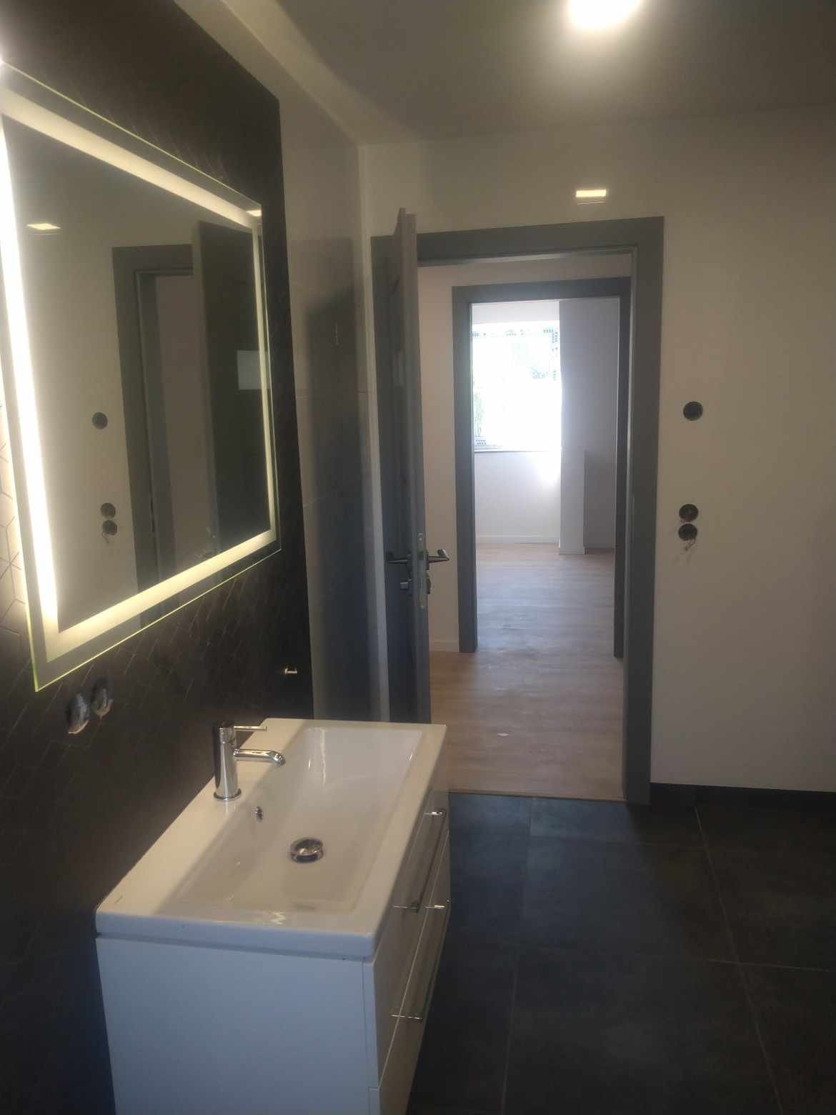 Nowoczesna łazienka z podświetlanym lustrem, białą umywalką na szafce i ciemną glazurą na ścianie, widok na otwarte drzwi do kolejnego pomieszczenia z jasną podłogą.