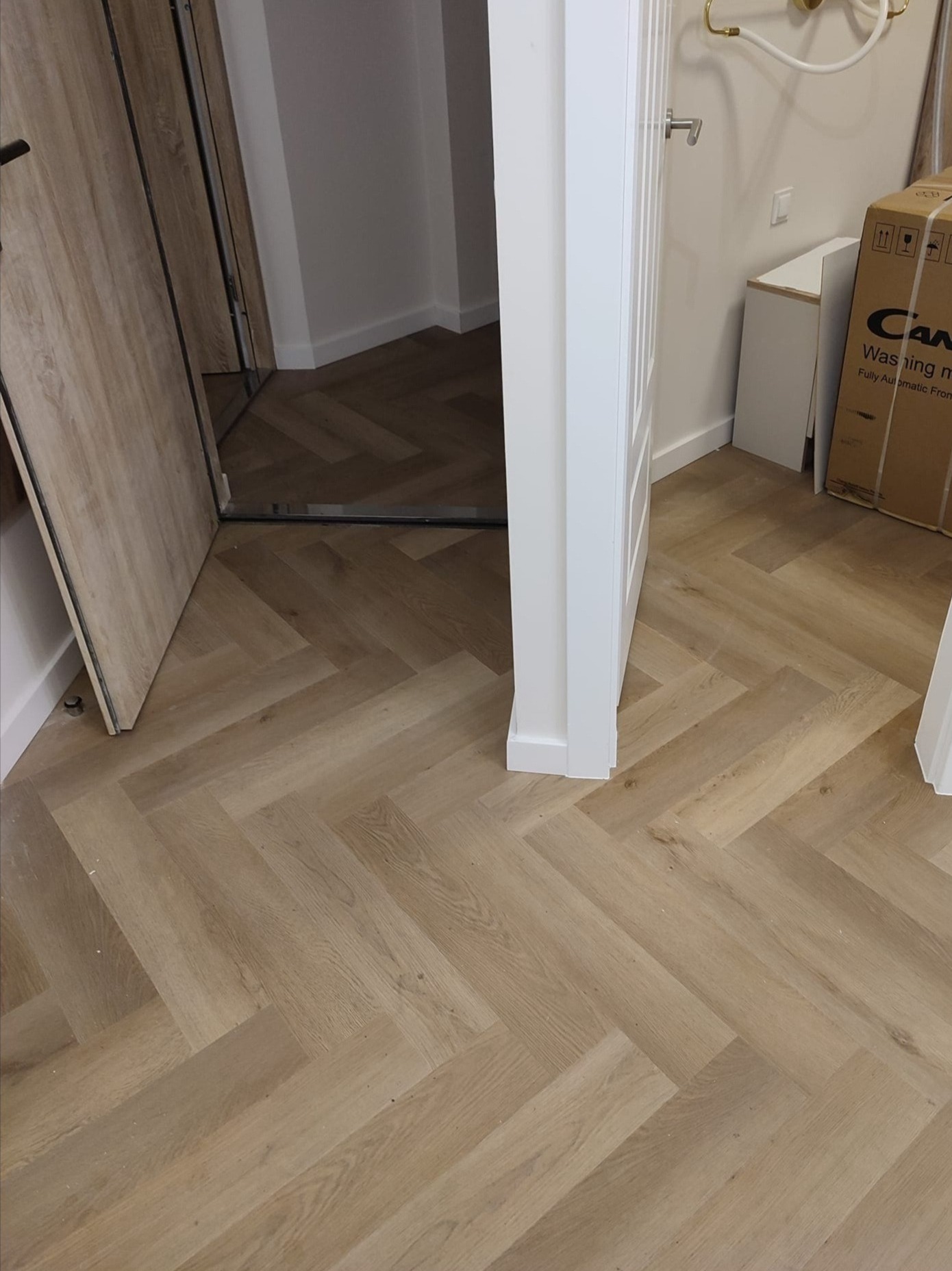 Ułożona podłoga z paneli imitujących drewno w jodełkę, widoczne fragmenty białych drzwi i futryny, w tle rozpakowane kartony i elementy wyposażenia łazienki.