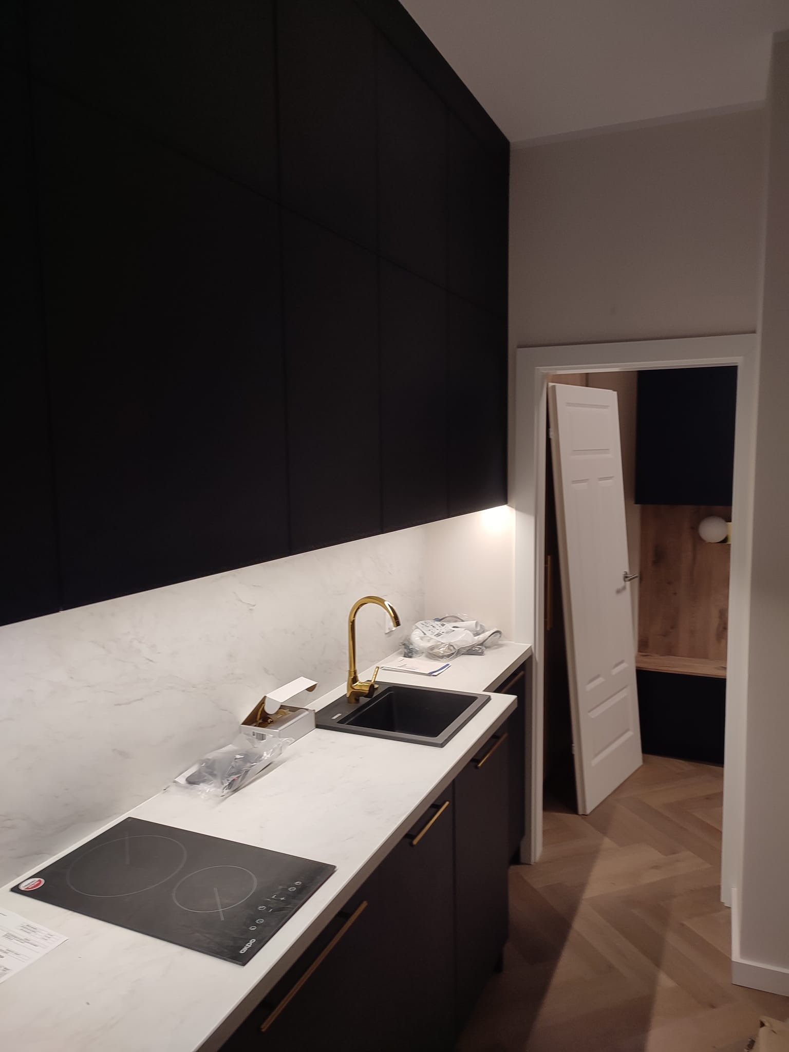 Nowoczesna kuchnia z czarnymi szafkami, marmurowym blatem, złotą armaturą i płytą indukcyjną, widok na otwarte drzwi do innego pomieszczenia z parkietem ułożonym w jodełkę.