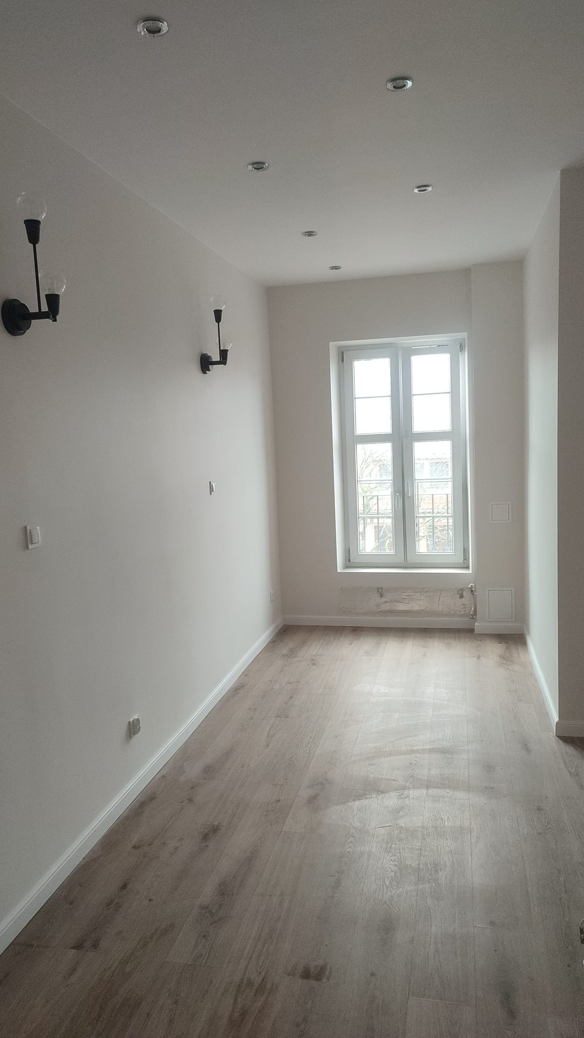 Jasne, puste pomieszczenie z drewnianą podłogą, białymi ścianami, oknem z widokiem na drzewa i nowoczesnymi czarnymi lampami ściennymi.