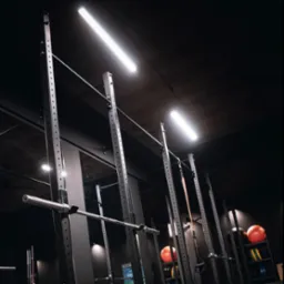 Wnętrze ciemnej siłowni z widocznymi stojakami na sztangi, obciążeniami i piłkami lekarskimi, oświetlone podłużnymi lampami sufitowymi.