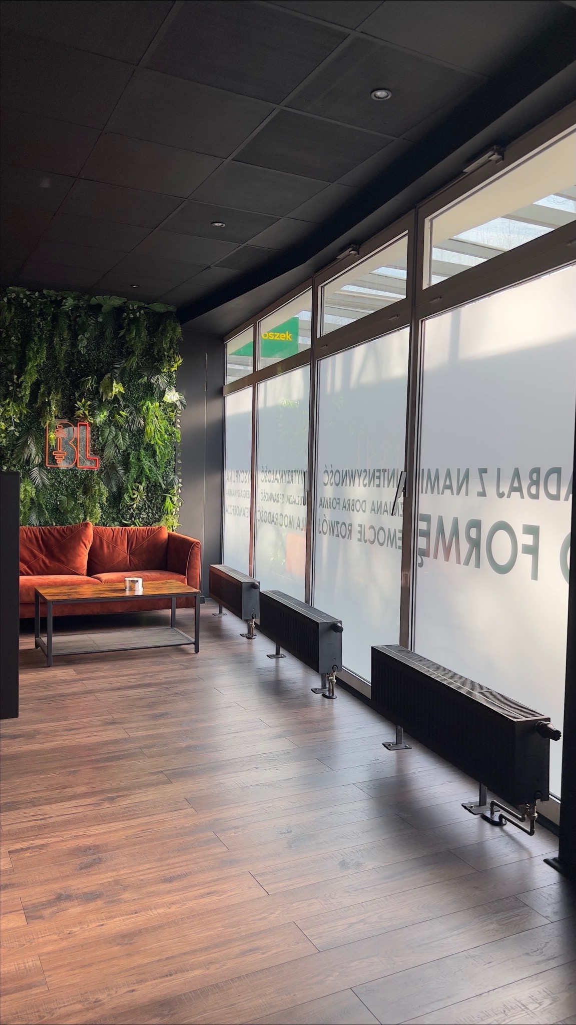 Nowoczesne wnętrze siłowni lub studia fitness z sofą, stolikiem kawowym, ścianą z roślin i neonowym logo 'BL' na ścianie. Na oknach widoczne napisy reklamowe.