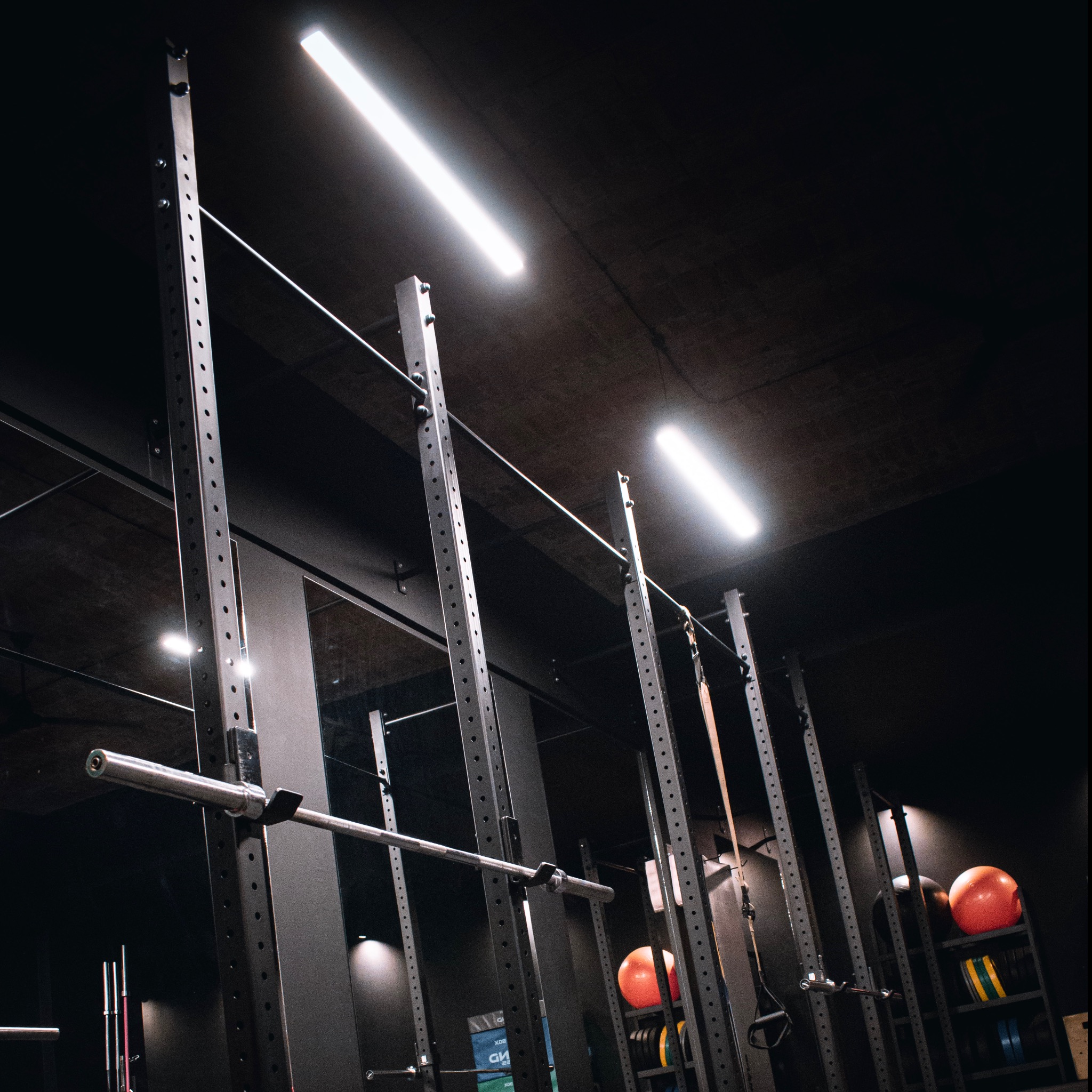 Wnętrze ciemnej siłowni z widocznymi stojakami na sztangi, obciążeniami i piłkami lekarskimi, oświetlone podłużnymi lampami sufitowymi.