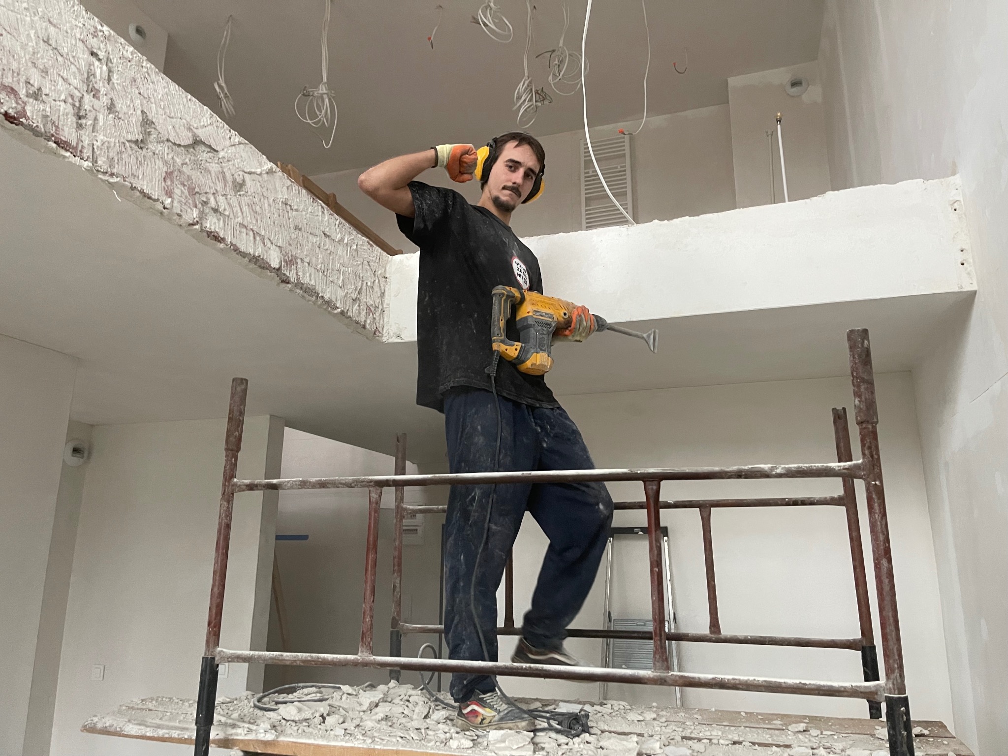 Mężczyzna z wąsami w słuchawkach ochronnych, stojący na rusztowaniu z młotem udarowym, w trakcie usuwania fragmentu ściany wewnętrznej, widoczne fragmenty gruzu i instalacji elektrycznej.
