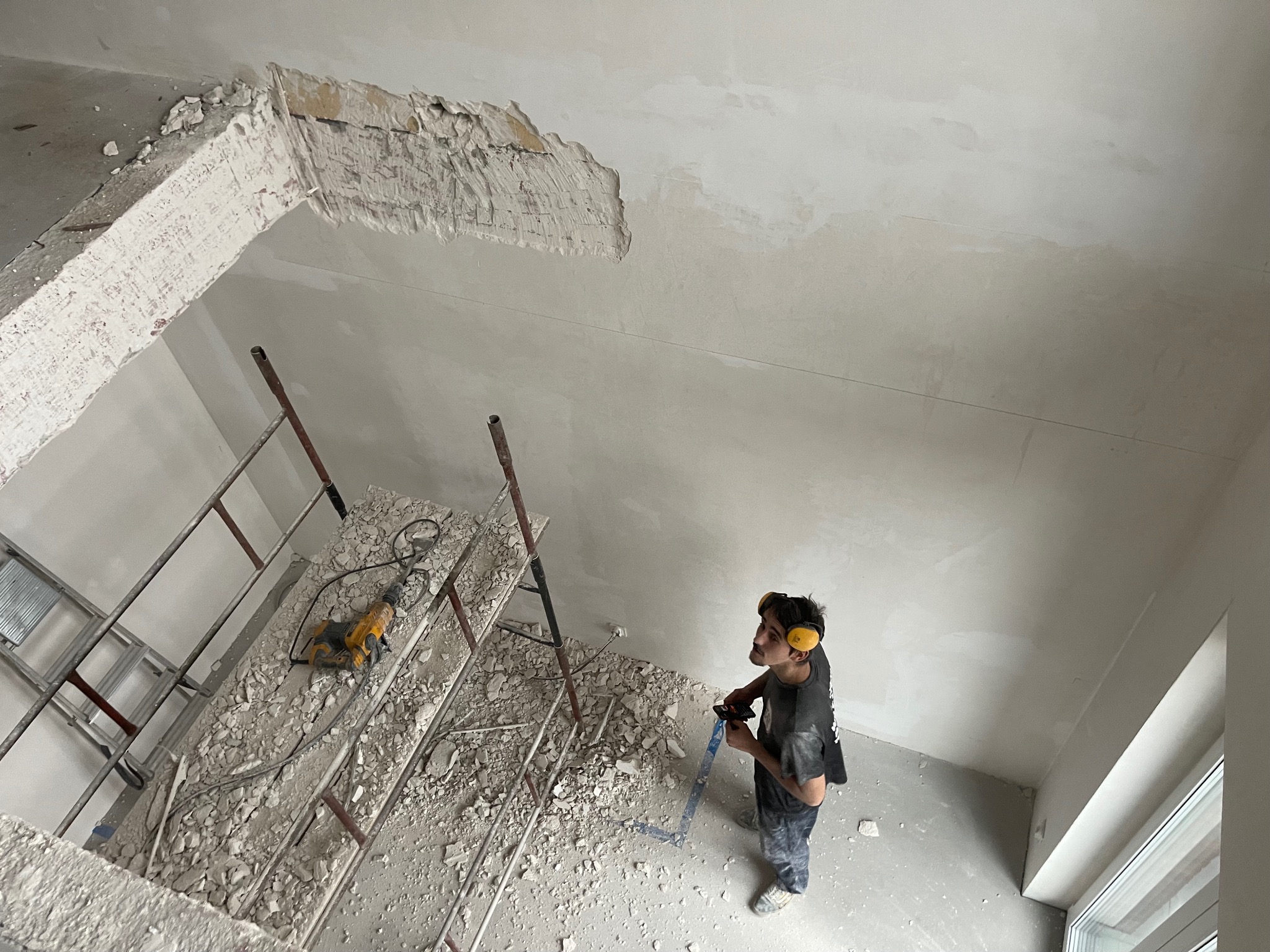 Pracownik w słuchawkach ochronnych, stojący na gruzowisku po usunięciu fragmentu sufitu w trakcie prac remontowych w Warszawie, spogląda w górę na otwór.