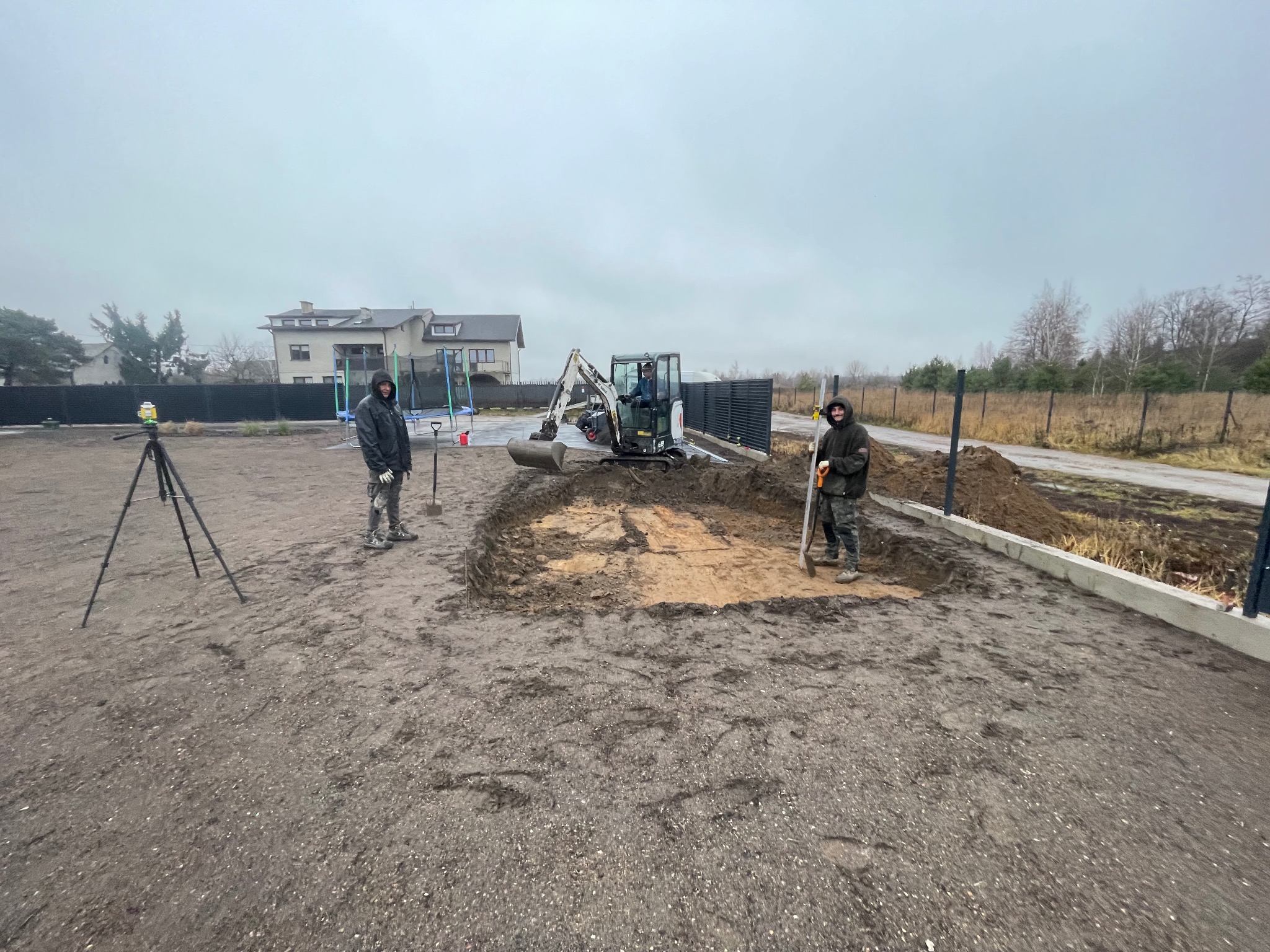Przygotowanie terenu pod budowę: mini koparka, pracownicy z łopatami i niwelator laserowy na statywie, wykopany kwadratowy dół z widoczną warstwą piasku.