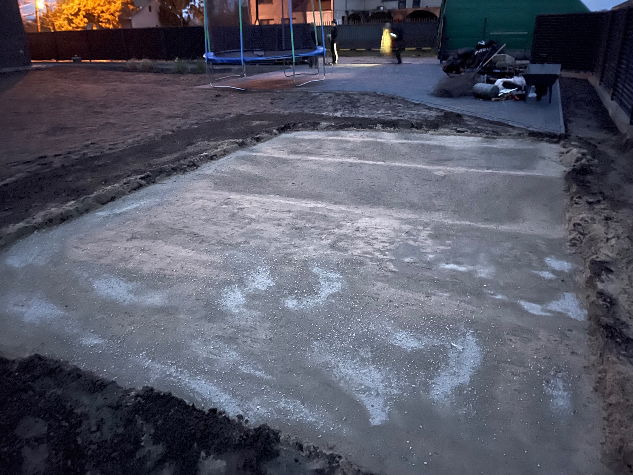 Wylana, świeża betonowa podstawa pod przyszłą konstrukcję na działce, widoczne ślady po poziomowaniu, ziemia wokół wykopu, w tle trampolina.