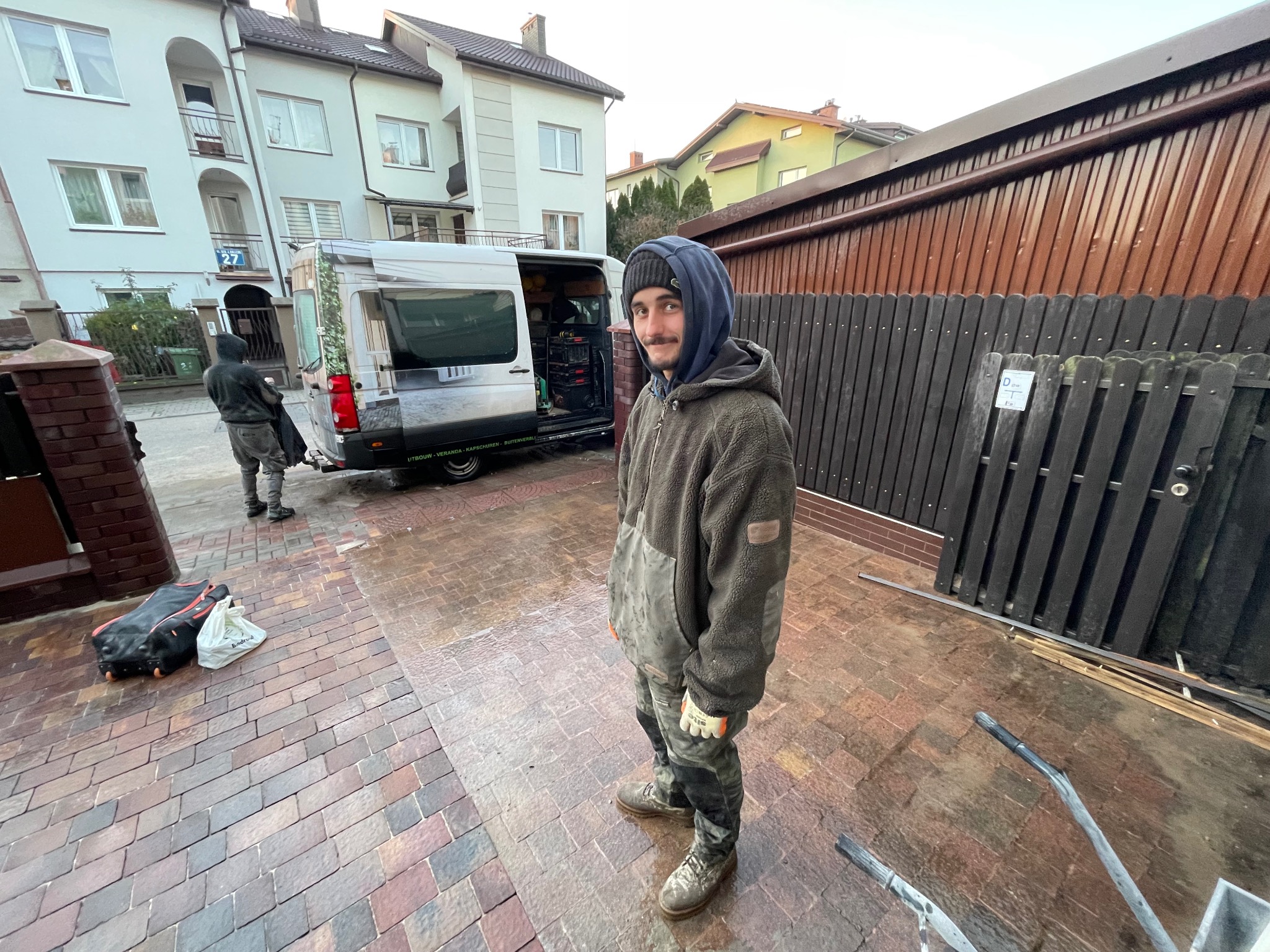 Pracownik w ubraniu roboczym stoi na posesji przed otwartym vanem z narzędziami, w tle budynki mieszkalne i drugi pracownik.