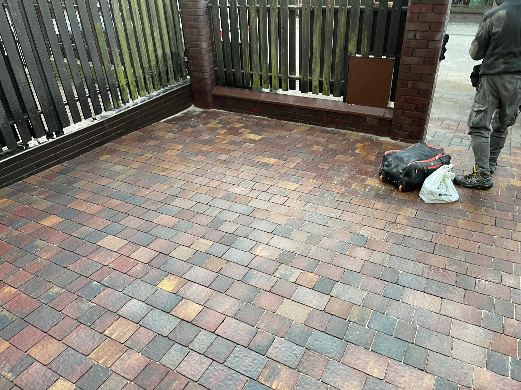 Ułożona kostka brukowa w odcieniach brązu i szarości, z widocznym fragmentem drewnianego ogrodzenia z ceglanymi słupkami, obok stoi torba narzędziowa i osoba w roboczym ubraniu.