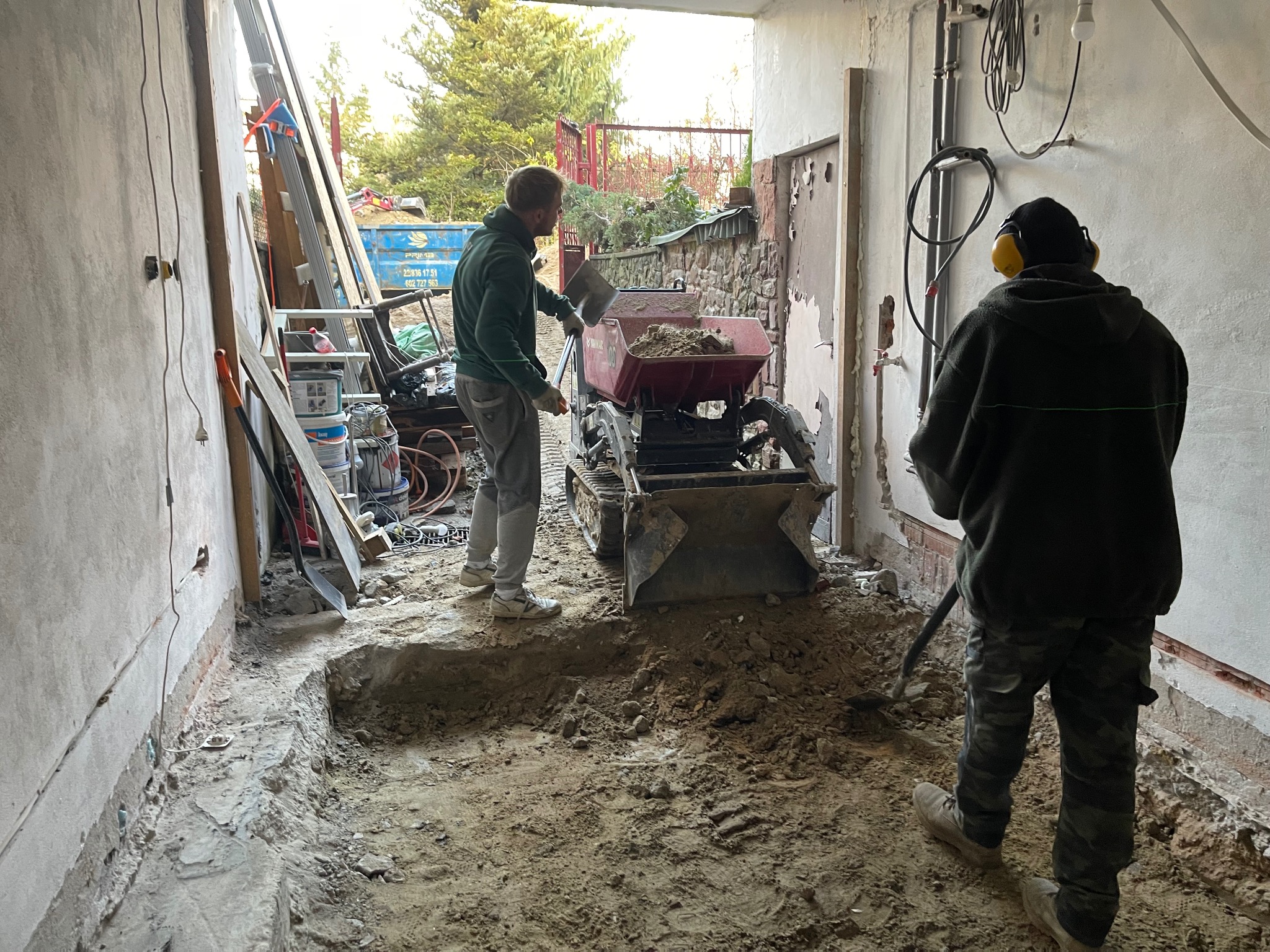 Dwóch robotników pracujących przy użyciu mini wywrotki i łopaty podczas robót ziemnych wewnątrz budynku, widoczne ślady wykopu w ziemi, ściany w stanie surowym.