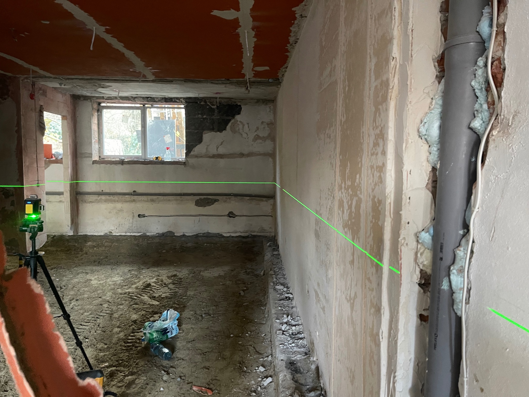 Wnętrze w trakcie remontu, z widocznymi śladami po usuniętych elementach, laser krzyżowy wyznacza poziom na ścianie, rura kanalizacyjna zabezpieczona pianką montażową.