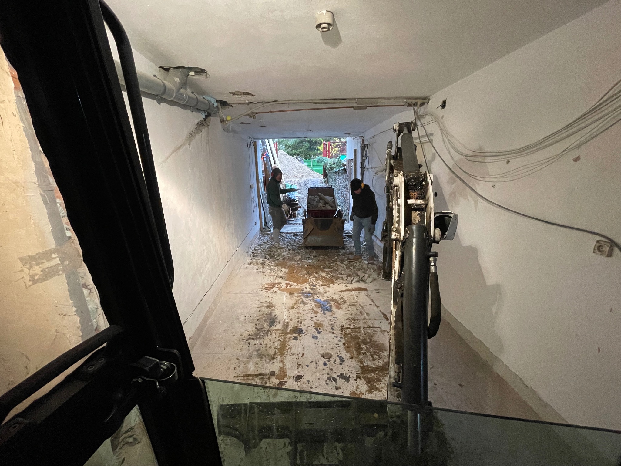 Wnętrze w trakcie prac: dwaj robotnicy ładują gruz do wózka, widok z kabiny maszyny budowlanej, rury pod sufitem
