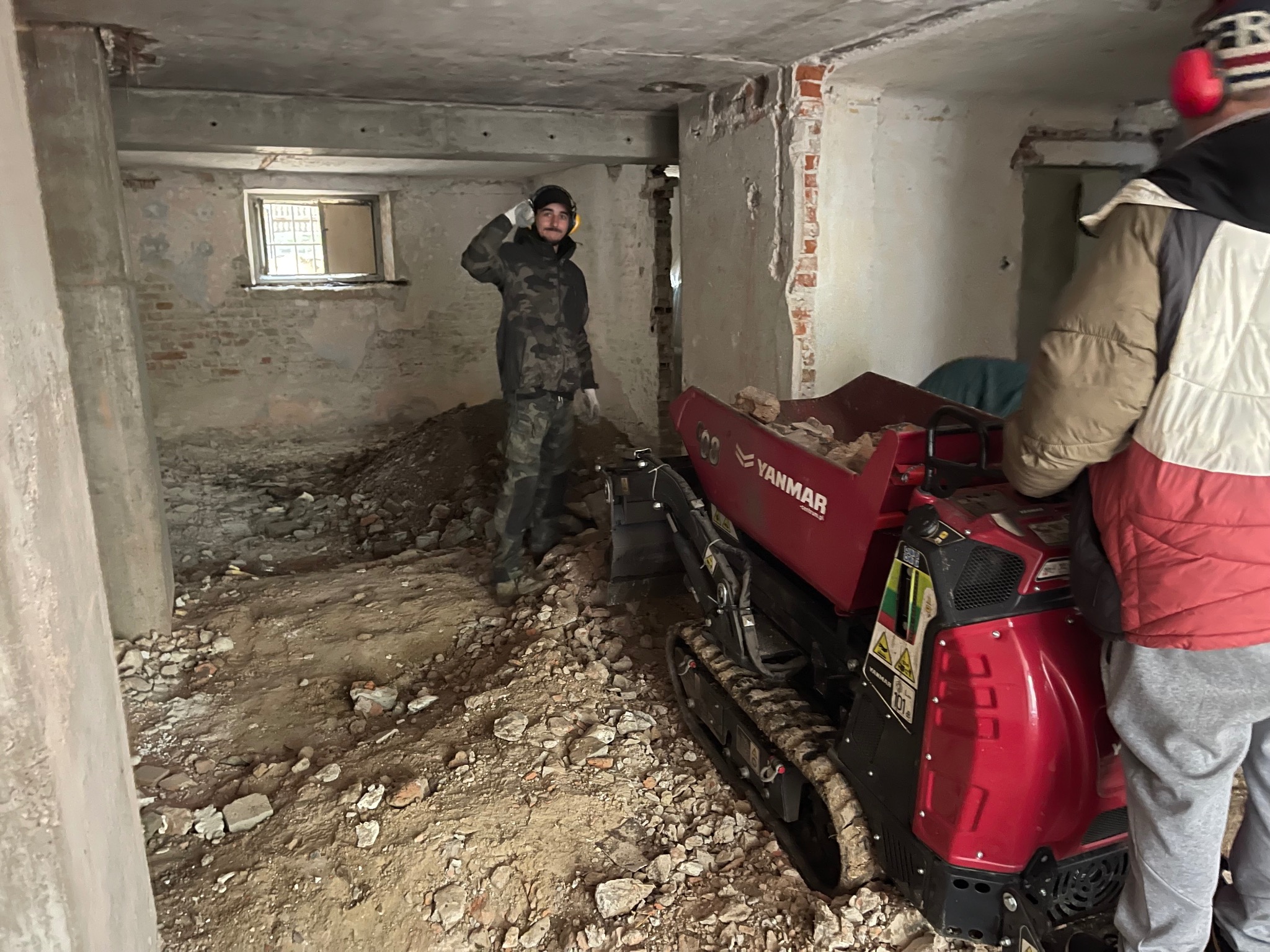 Wnętrze piwnicy w trakcie prac, z gruzem na podłodze, pracownikiem w stroju roboczym i minikoparką Yanmar.