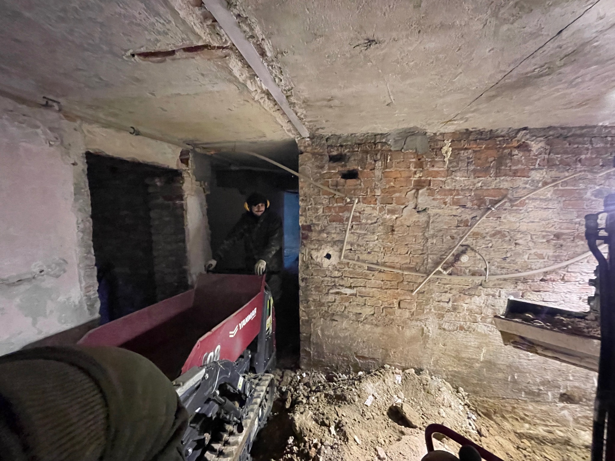 Pracownik w słuchawkach ochronnych obsługuje mini-wywrotkę Yanmar w trakcie prac wewnątrz budynku z odsłoniętą cegłą i częściowo usuniętym tynkiem, widoczne gruz i resztki materiałów budowlanych.