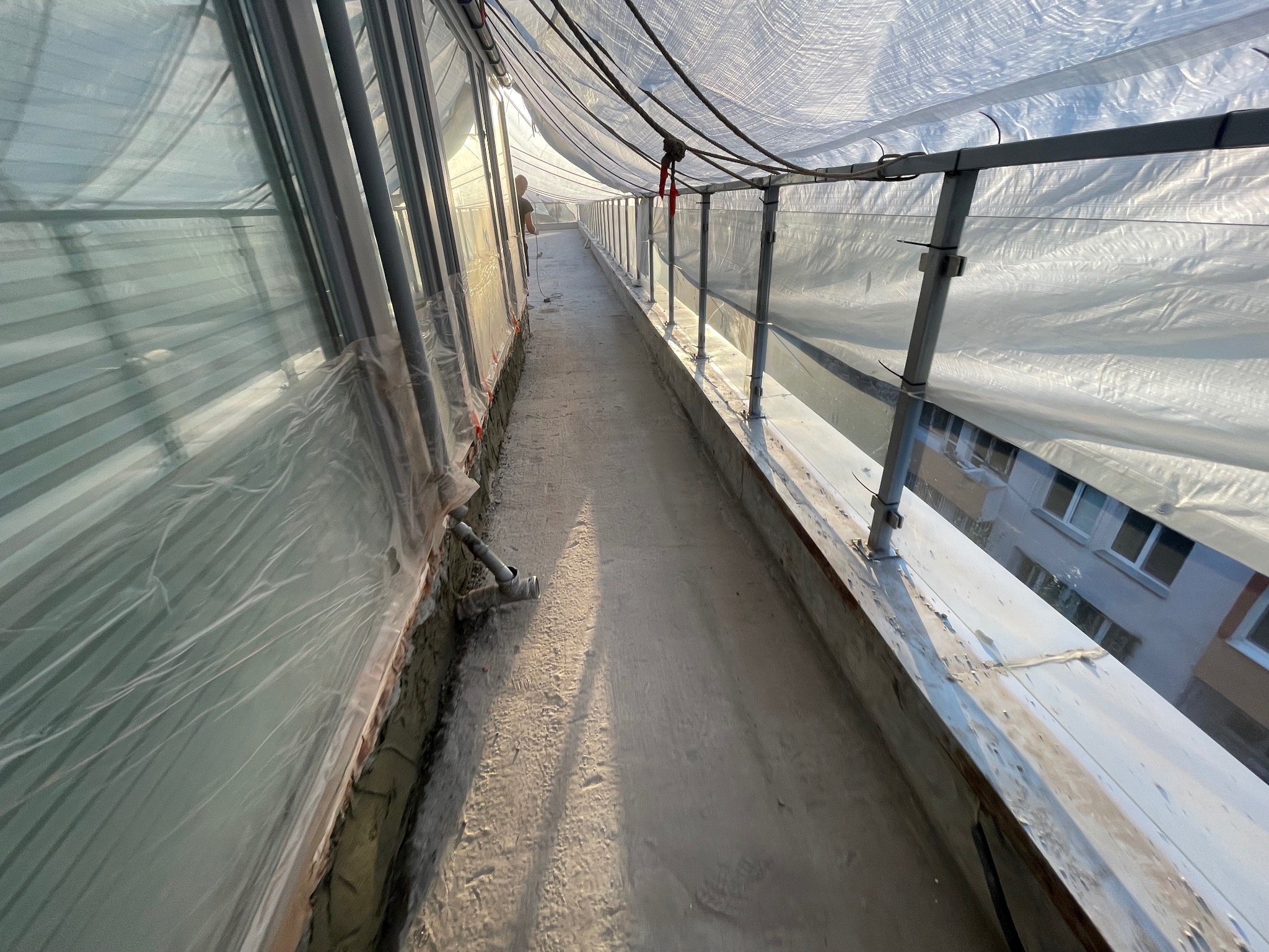 Długi balkon zabezpieczony folią i metalowymi barierkami w trakcie prac budowlanych, widoczny pracownik w oddali.