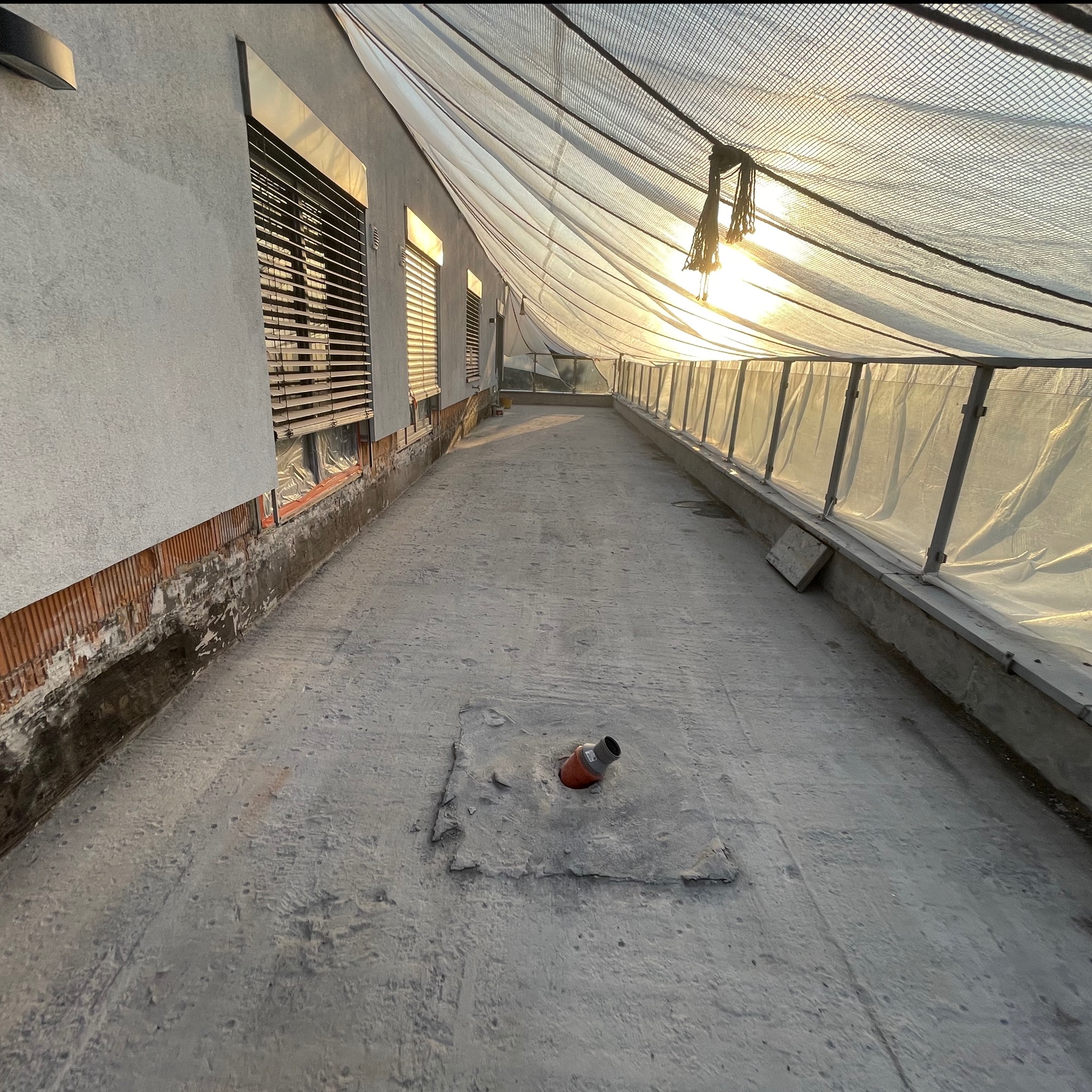 Przestrzeń w trakcie prac budowlanych z odsłoniętymi cegłami, zabezpieczonymi oknami i rurą wystającą z posadzki pod osłoną siatki.