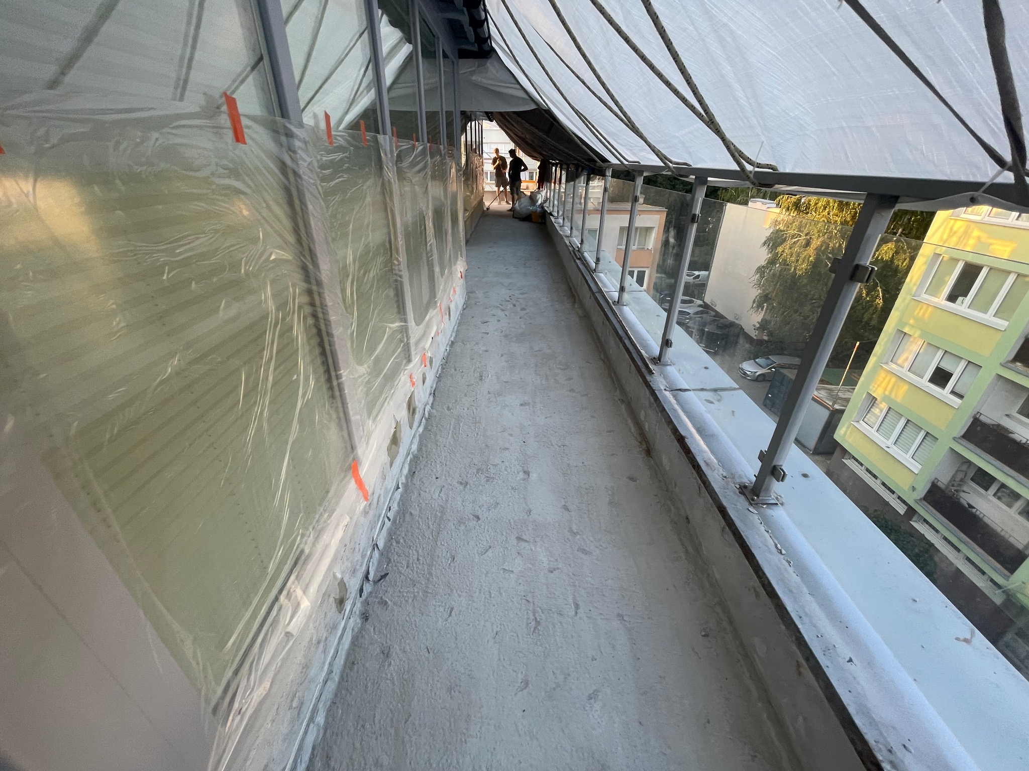 Balkon w trakcie remontu, osłonięty folią i plandeką, widoczni pracownicy w oddali.