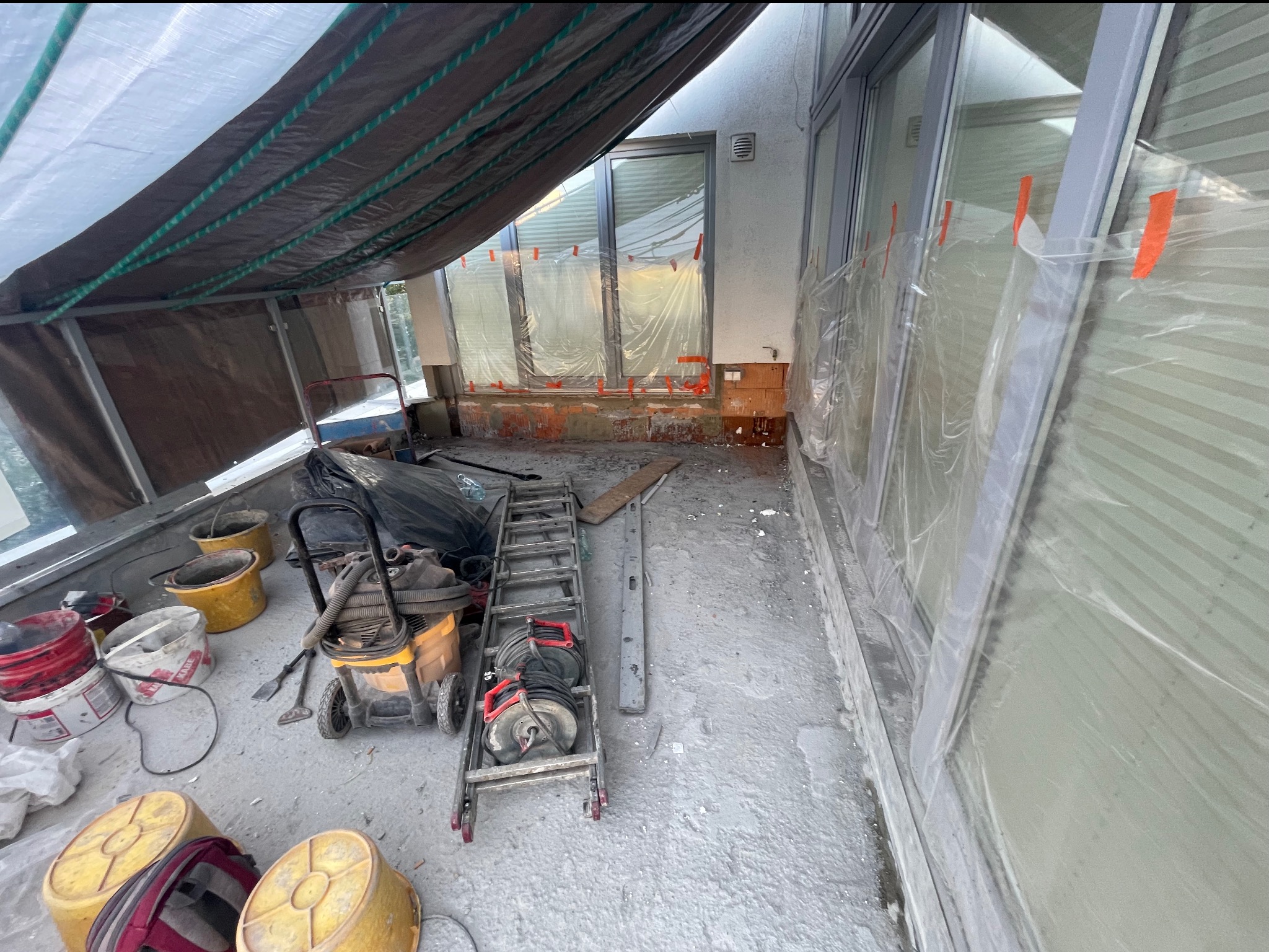 Przygotowania do prac budowlanych na balkonie: zabezpieczone okna folią, rozstawione narzędzia, wiaderka, odkurzacz przemysłowy i drabina pod zadaszeniem.