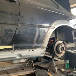 Czarny samochód na podnośniku w warsztacie, widoczne prace blacharskie w toku: spawanie i dopasowywanie nowego progu, odsłonięte zawieszenie z czerwoną sprężyną, narzędzia leżące obok koła.