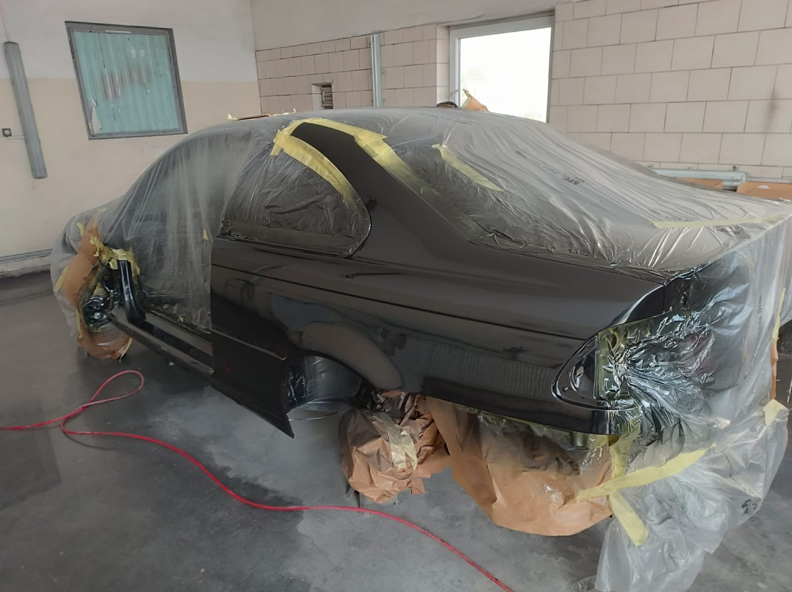 Czarny samochód w trakcie lakierowania w warsztacie, zabezpieczony folią i taśmą maskującą, widoczny przewód lakierniczy na podłodze.