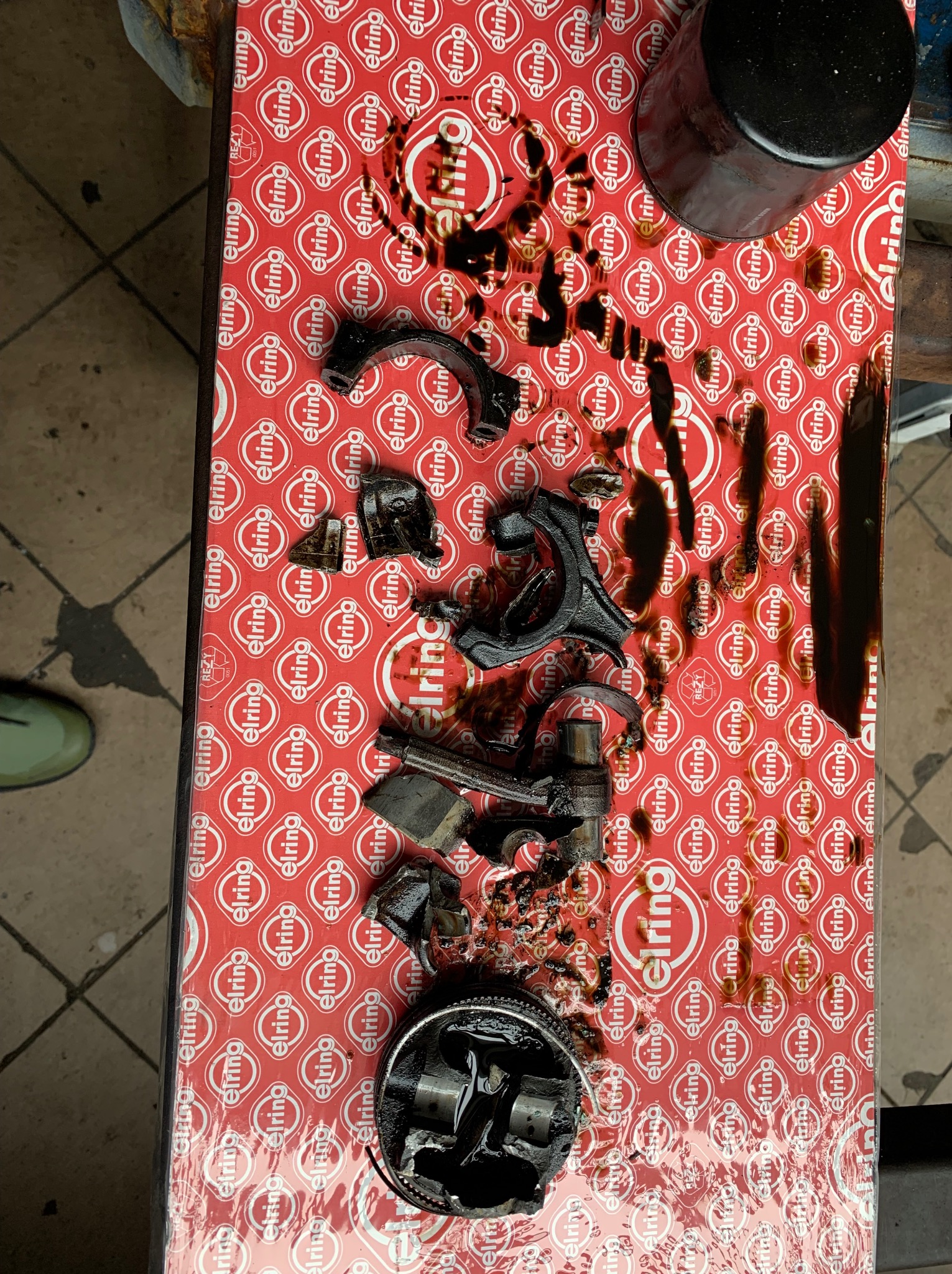 Rozłożony silnik samochodowy z wyciekającym olejem na kartonie Elring, widoczne elementy tłoka, korbowody i filtr oleju.