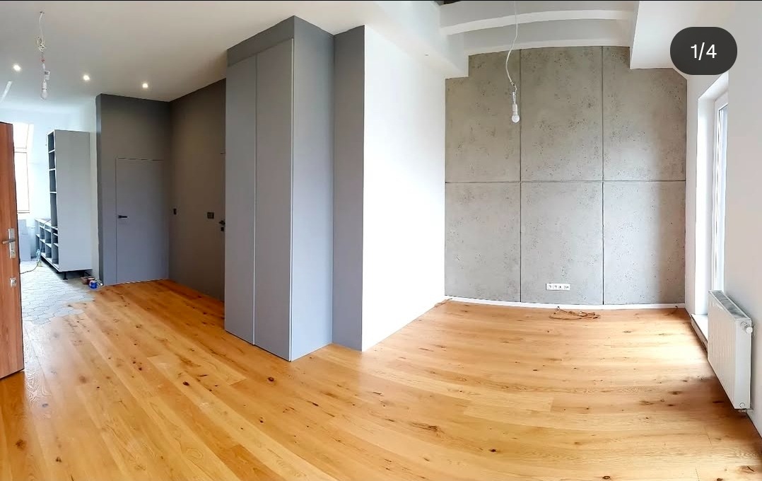 Jasne drewniane podłogi w przestronnym wnętrzu z szarą zabudową i betonową ścianą. Widoczne detale wykończenia, surowy styl, minimalistyczny design.