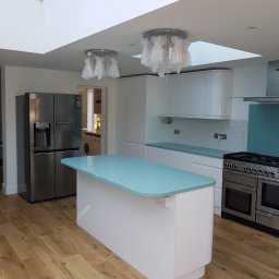 WIDAM Adam Wicha Usługi Remontowo-Budowlane - Jasna, przestronna kuchnia z wyspą o turkusowym blacie, białe szafki z zaokrąglonymi krawędziami, duża lodówka side-by-side, kuchenka z piekarnikiem i okapem, drewniana podłoga, lampy w folii...