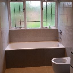 WIDAM Adam Wicha Usługi Remontowo-Budowlane - Wykończona łazienka z wanną obudowaną płytkami, wiszącą toaletą i oknem z kratownicą, wykończona beżowymi płytkami podłogowymi i ściennymi.